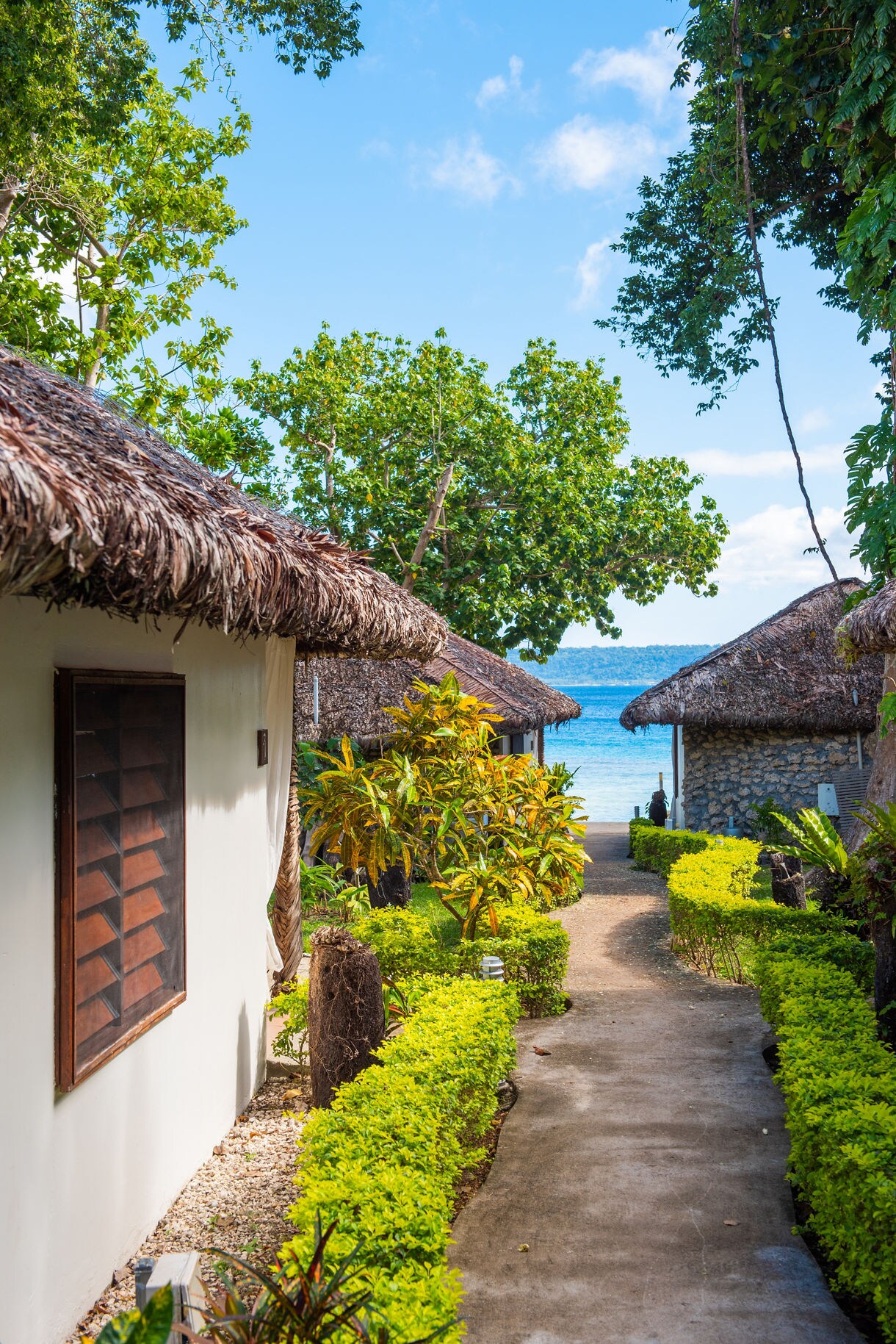 A narrow walkway lined with bright green shrubs leads between thatched-roof bungalows toward a blue ocean, framed by tall tropical trees under a clear sky.