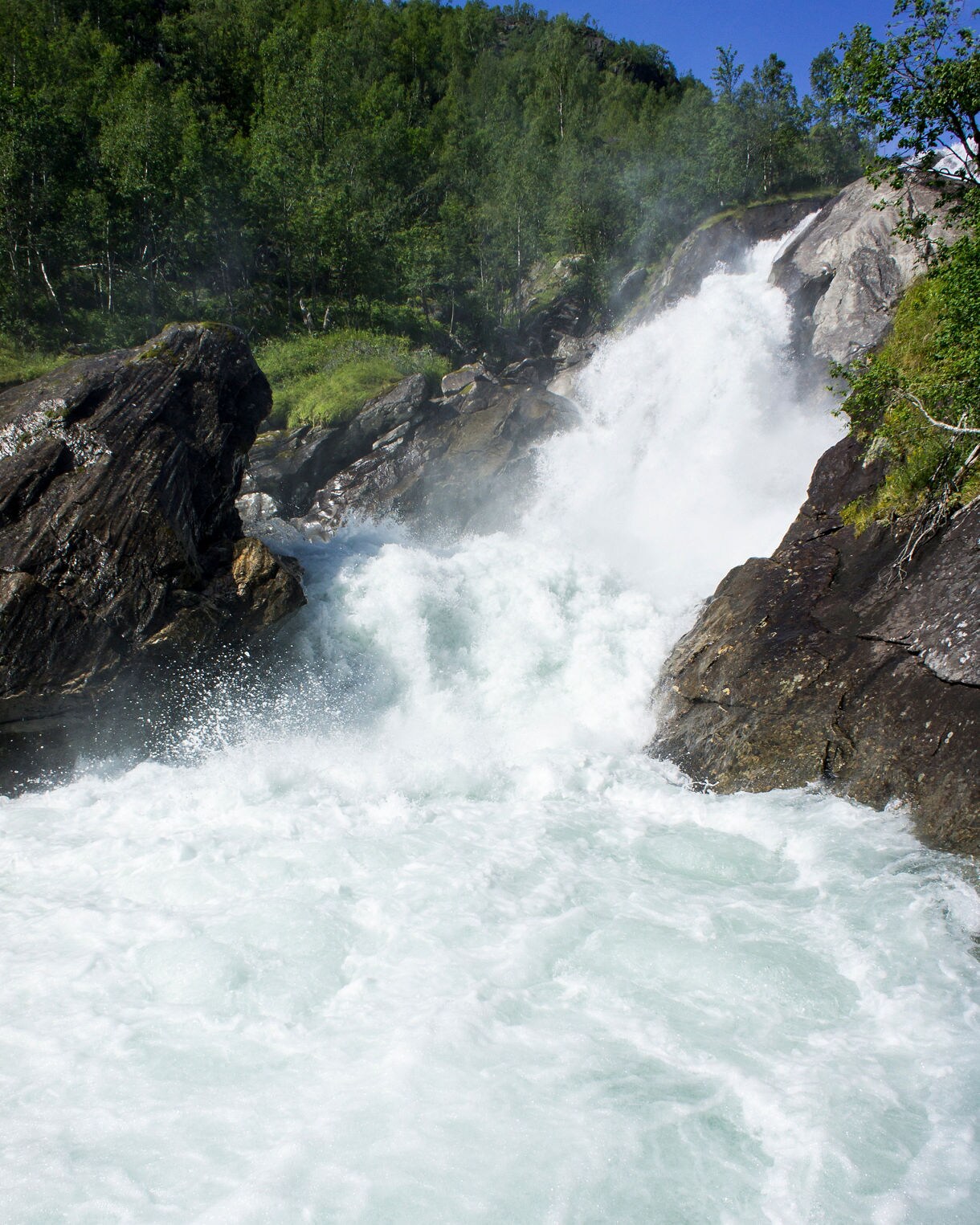 Forceful waterfall and rapids in Mørkridsdalen Valley near Skjolden, Norway, with foaming white water rushing between rocky cliffs surrounded by green forest.