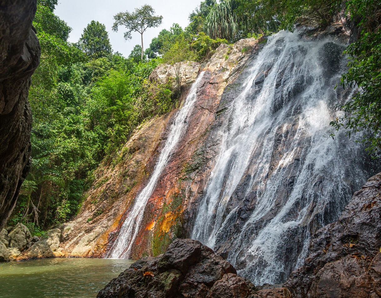 A tall waterfall flows over reddish and dark rock into a calm basin surrounded by dense green forest.