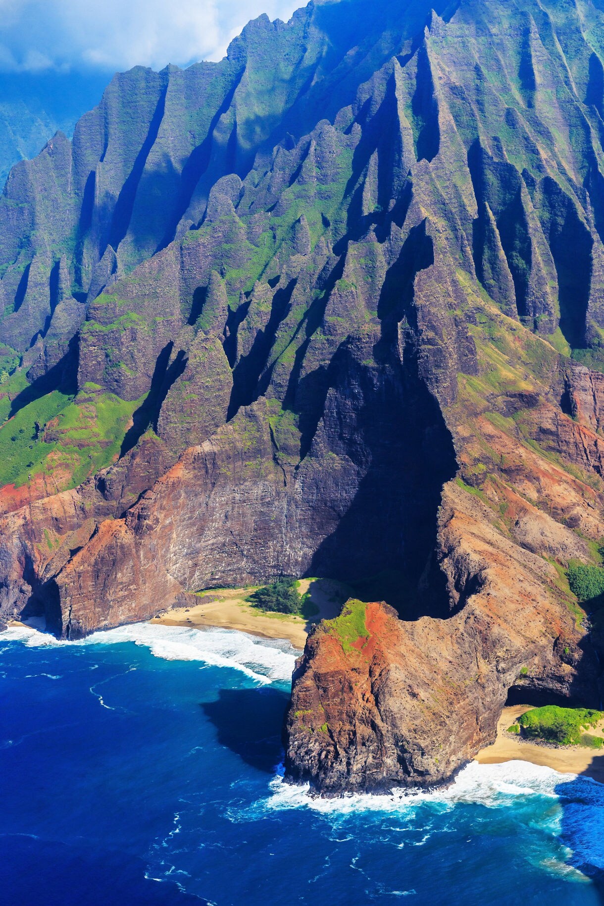 Aerial view of the Na Pali Coast showing steep green cliffs, rugged rock formations and golden beaches meeting deep blue ocean waves.