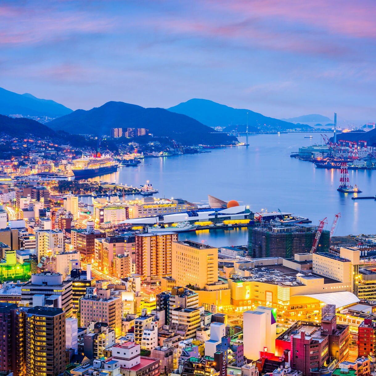 Elevated view of Nagasaki at twilight with illuminated buildings, busy harbor cranes, docked ships and calm blue water surrounded by dark mountains.