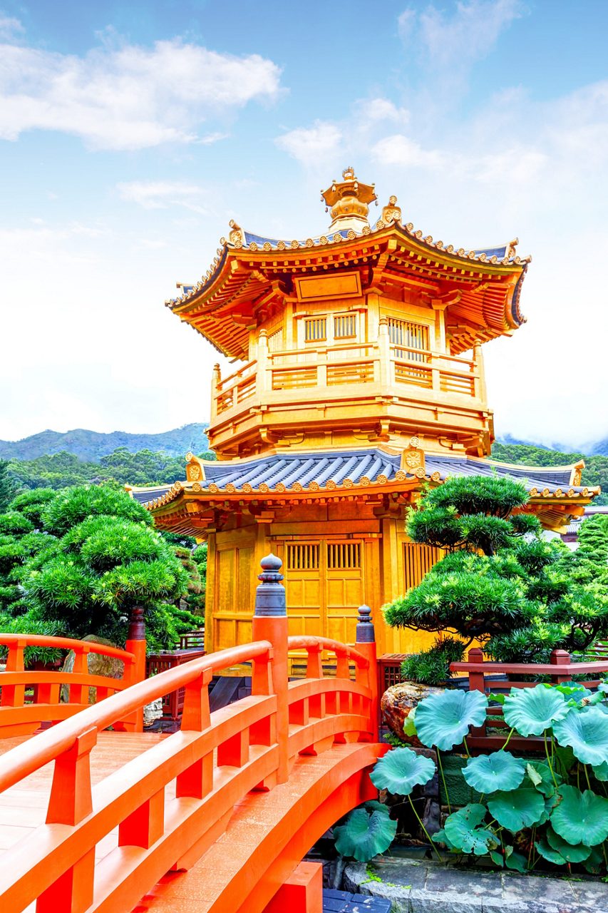 Golden pavilion at Nan Lian Garden in Hong Kong with a red wooden bridge, lotus plants and manicured trees under a bright blue sky.