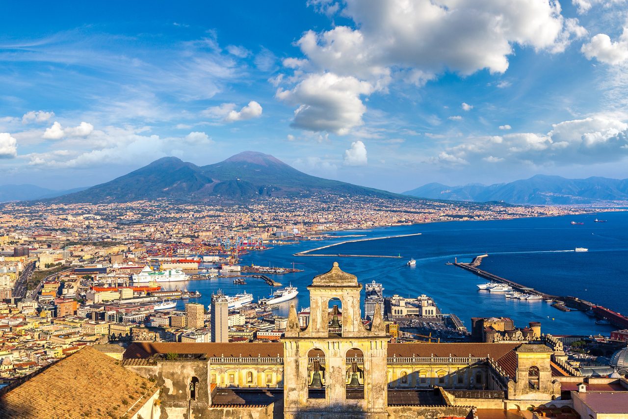 Panoramic view of Naples featuring historic buildings, a bustling harbor and Mount Vesuvius overlooking the coastline and blue Mediterranean Sea.