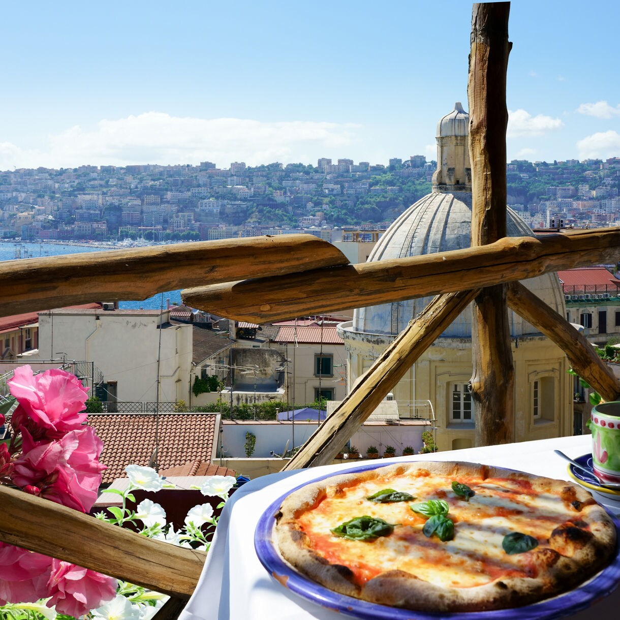Margherita pizza on a table beside an espresso cup, overlooking Naples rooftops, a church dome and the bay beyond.