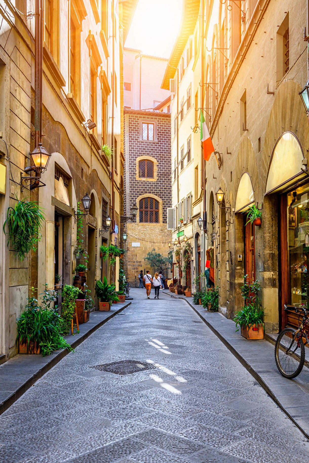 A narrow cobblestone street in Florence with old stone buildings, hanging lanterns and potted plants, a bicycle leaning against a wall and a couple walking in the distance.