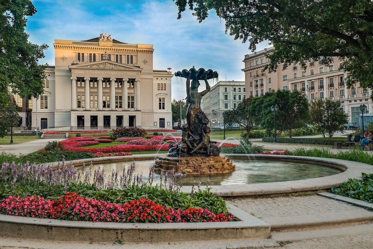 The Latvian National Opera House in Riga, a neoclassical building with tall columns and ornate pediment, seen from a park with a fountain and colorful flowerbeds in the foreground.