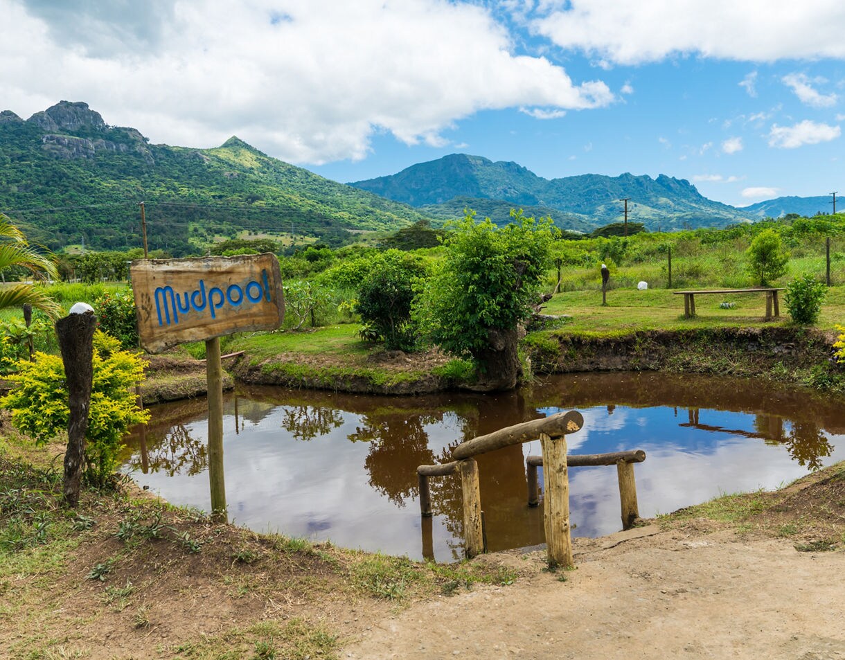 Rustic mud pool in a green valley in Fiji with a wooden sign, small pond and mountains rising in the background under a partly cloudy sky.