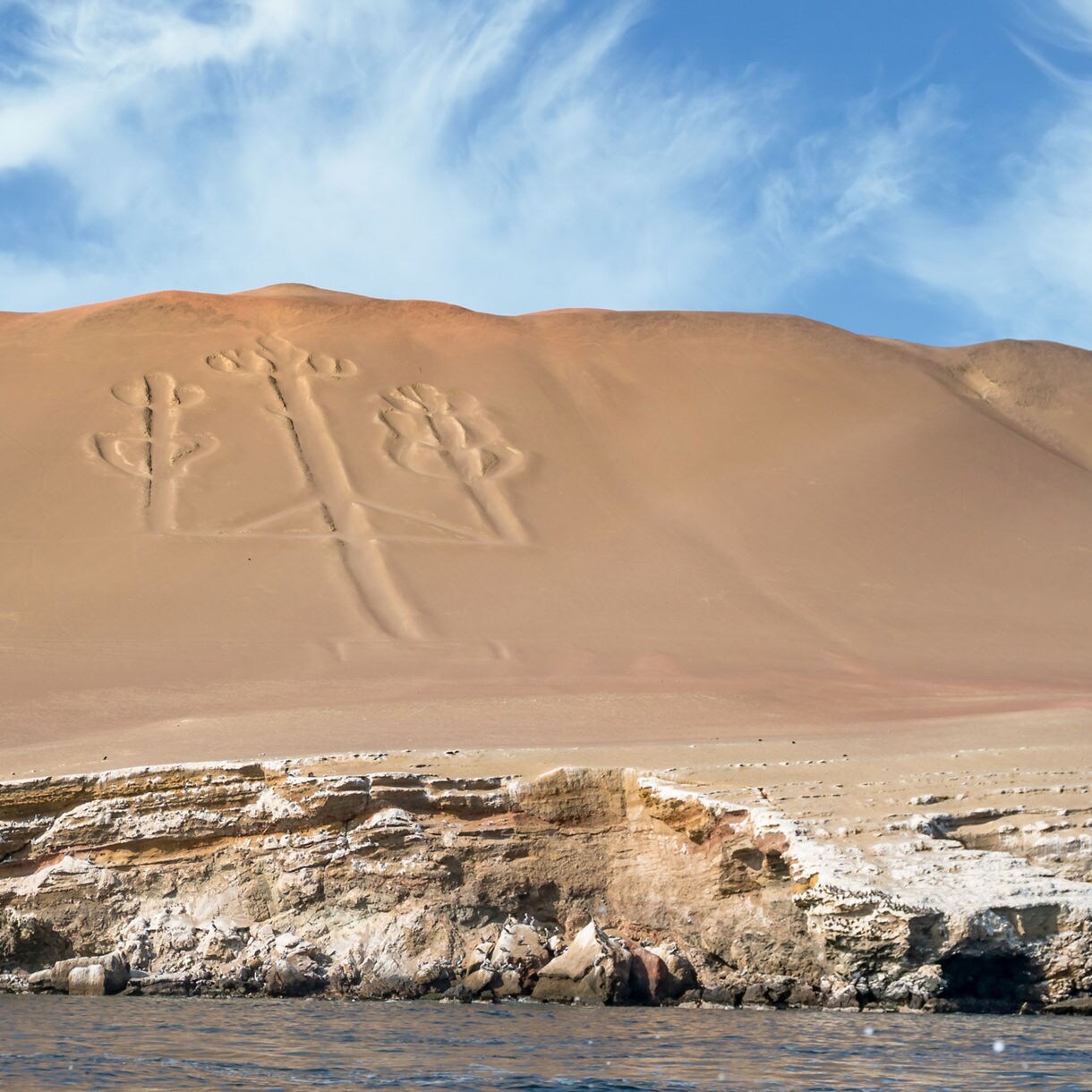 Large ancient geoglyph shaped like a three-branched candelabra carved into a sandy hillside above rocky coastal cliffs and calm ocean water.
