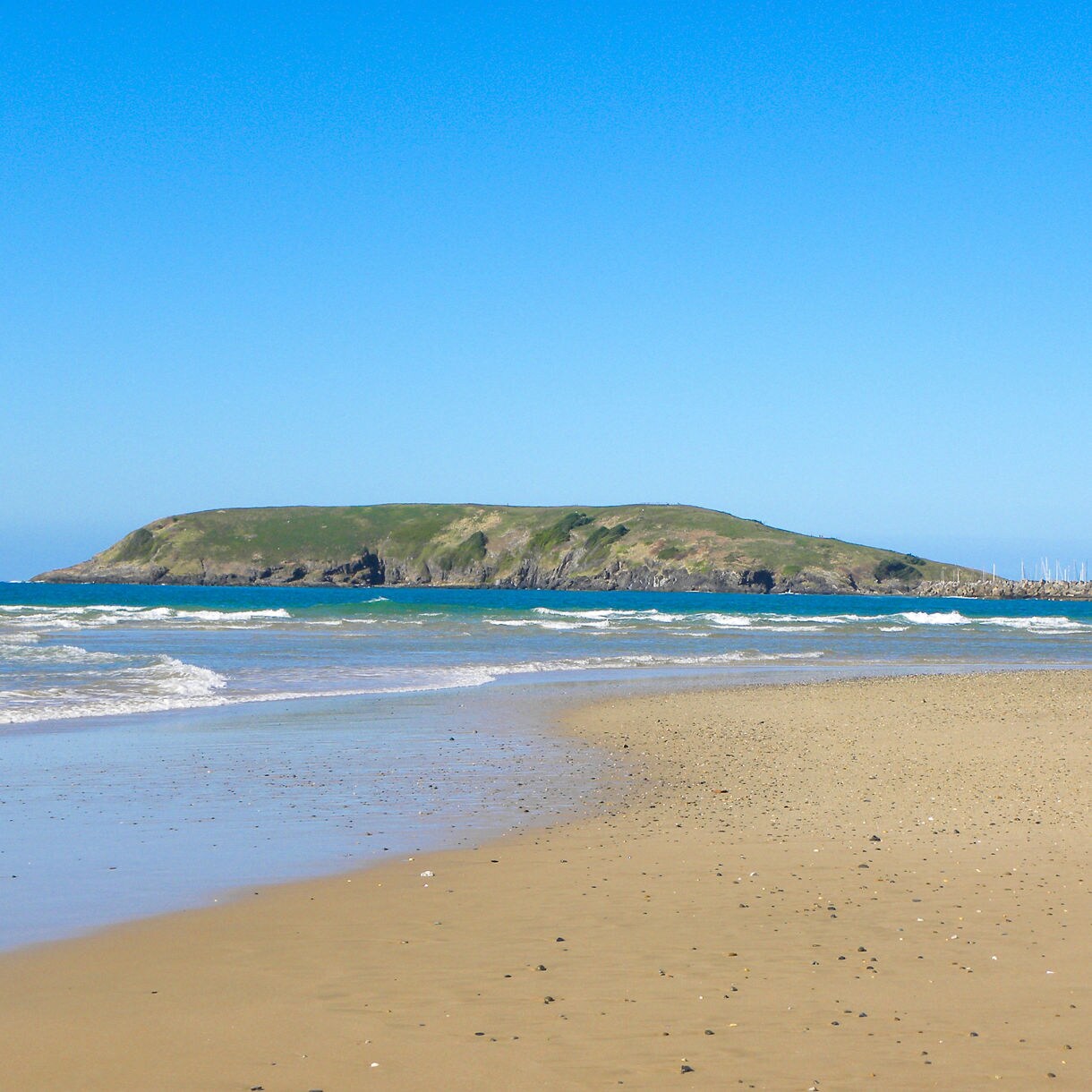 Wide sandy beach with small waves rolling in, a green headland in the distance and clear blue sky overhead at Nelson Bay.