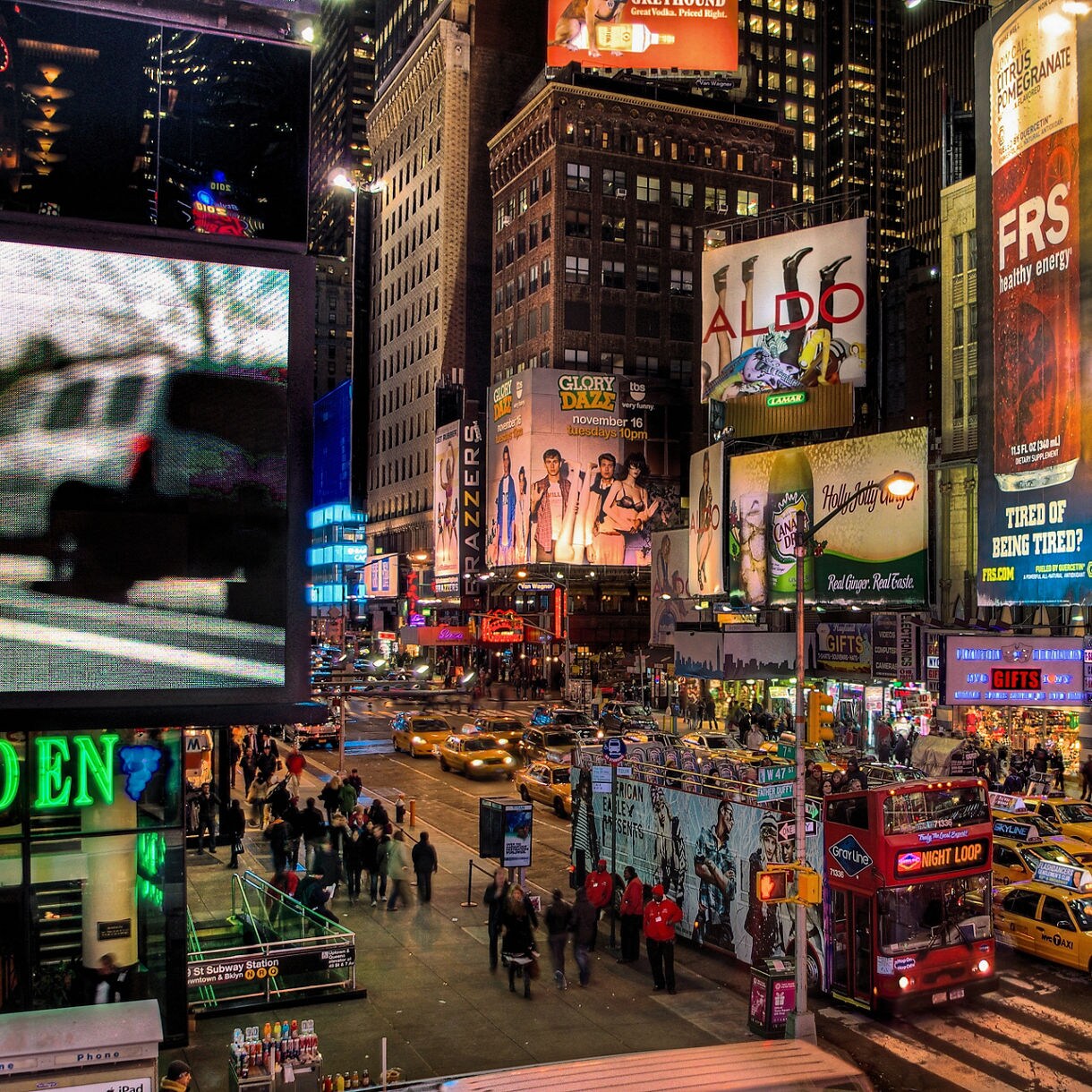 Busy nighttime scene in the Broadway theater district with bright billboards, yellow taxis, crowds crossing the street and illuminated signs for popular shows.