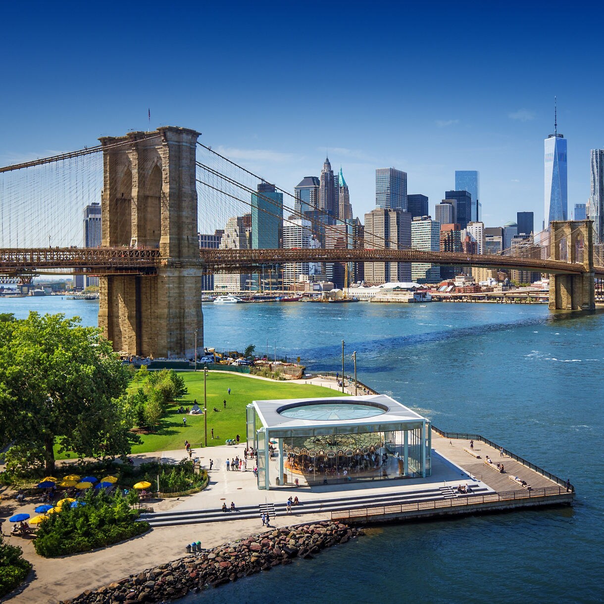 A wide aerial view of the Brooklyn Bridge spanning the East River with Manhattan’s skyscrapers in the background, a waterfront park and glass-enclosed carousel in the foreground and a ferry crossing the water.