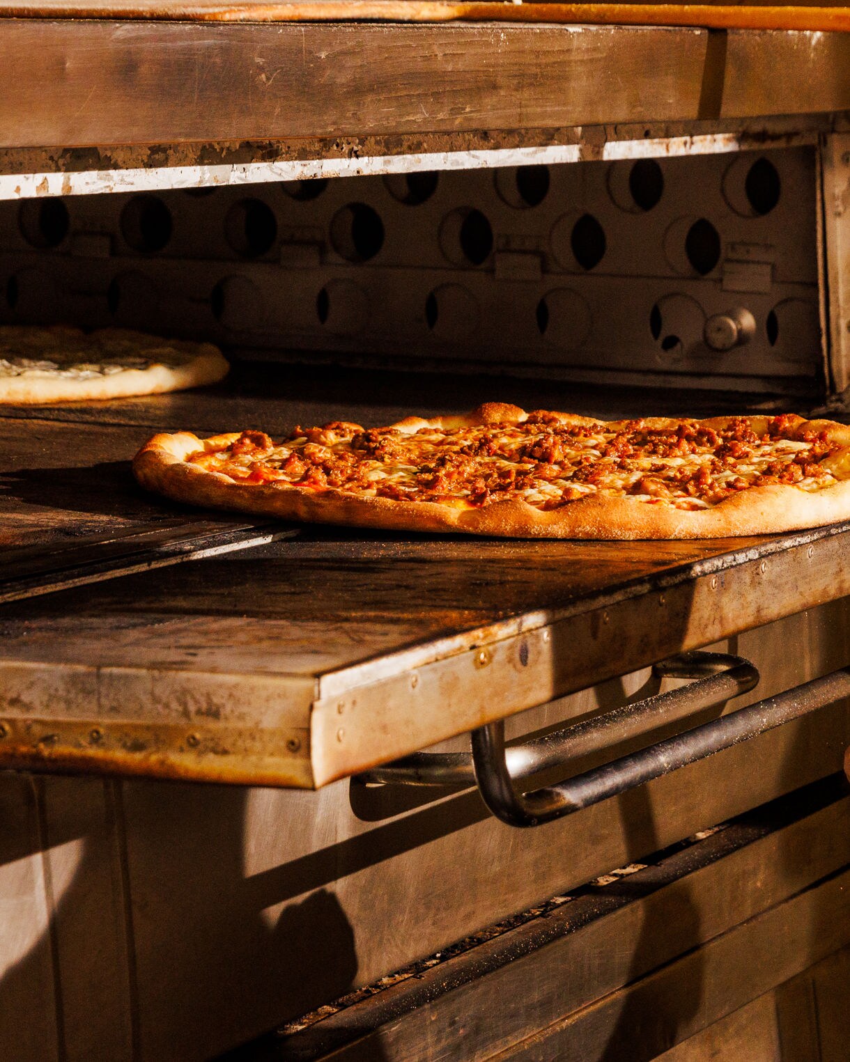 Close-up of a fresh pizza being pulled from a commercial oven with a wooden peel, showing a golden crust and melted toppings.