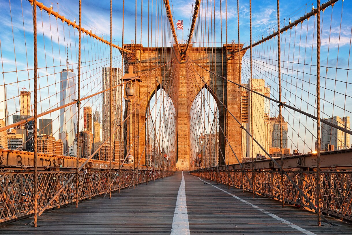 View from the pedestrian walkway of the Brooklyn Bridge at sunrise, showing its lattice of cables, stone towers and the Manhattan skyline glowing in warm early light.