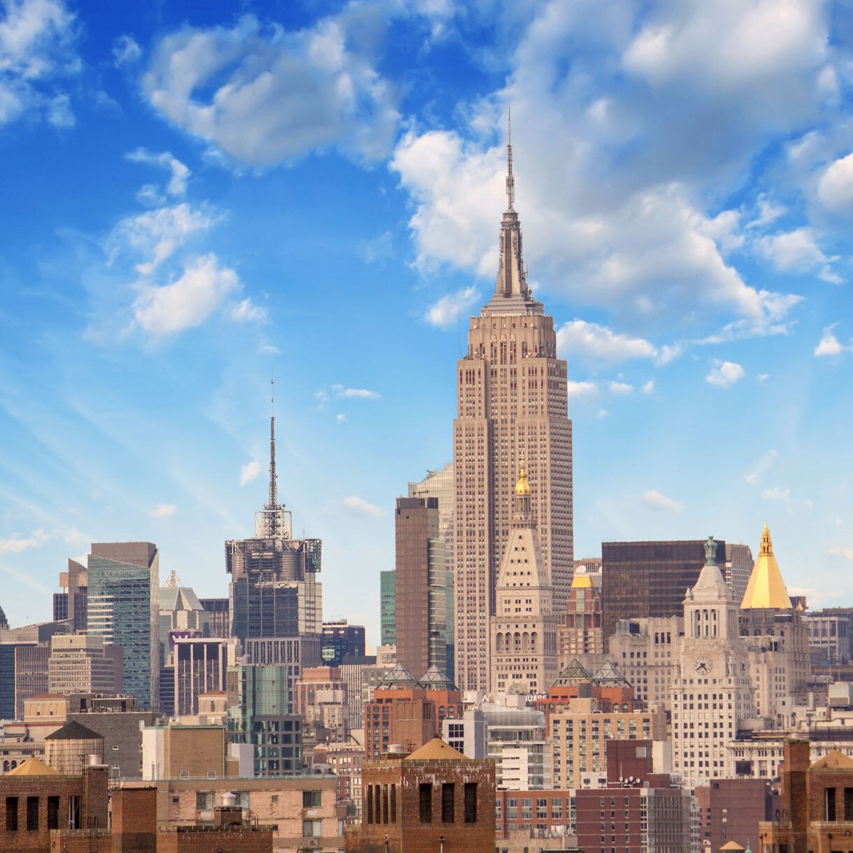 Panoramic view of midtown Manhattan with the Empire State Building rising above surrounding skyscrapers under a vivid sky filled with soft clouds.