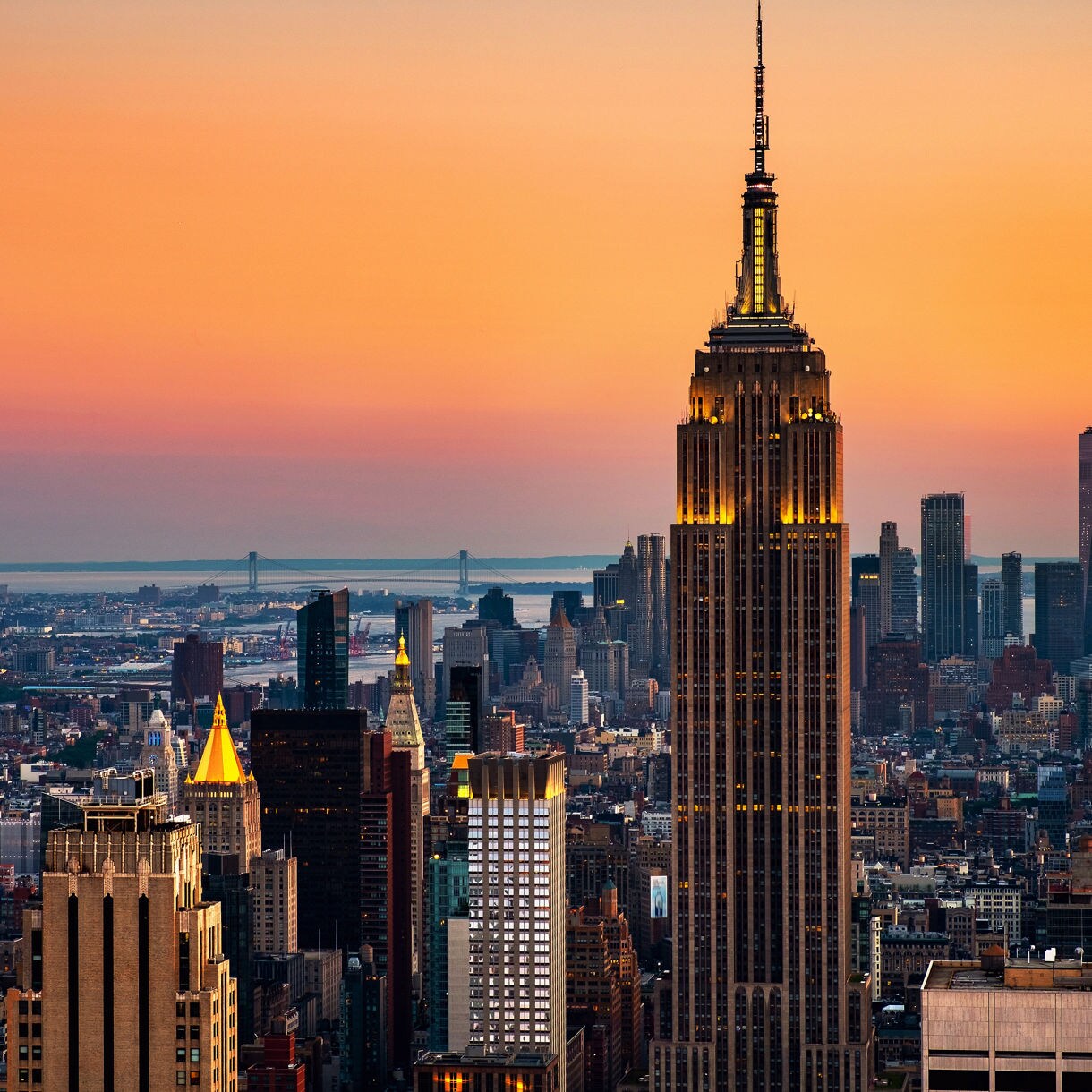 Aerial view of the Empire State Building at dusk with warm lights on its tower, surrounded by Manhattan’s skyline and an orange gradient sunset stretching across the horizon.