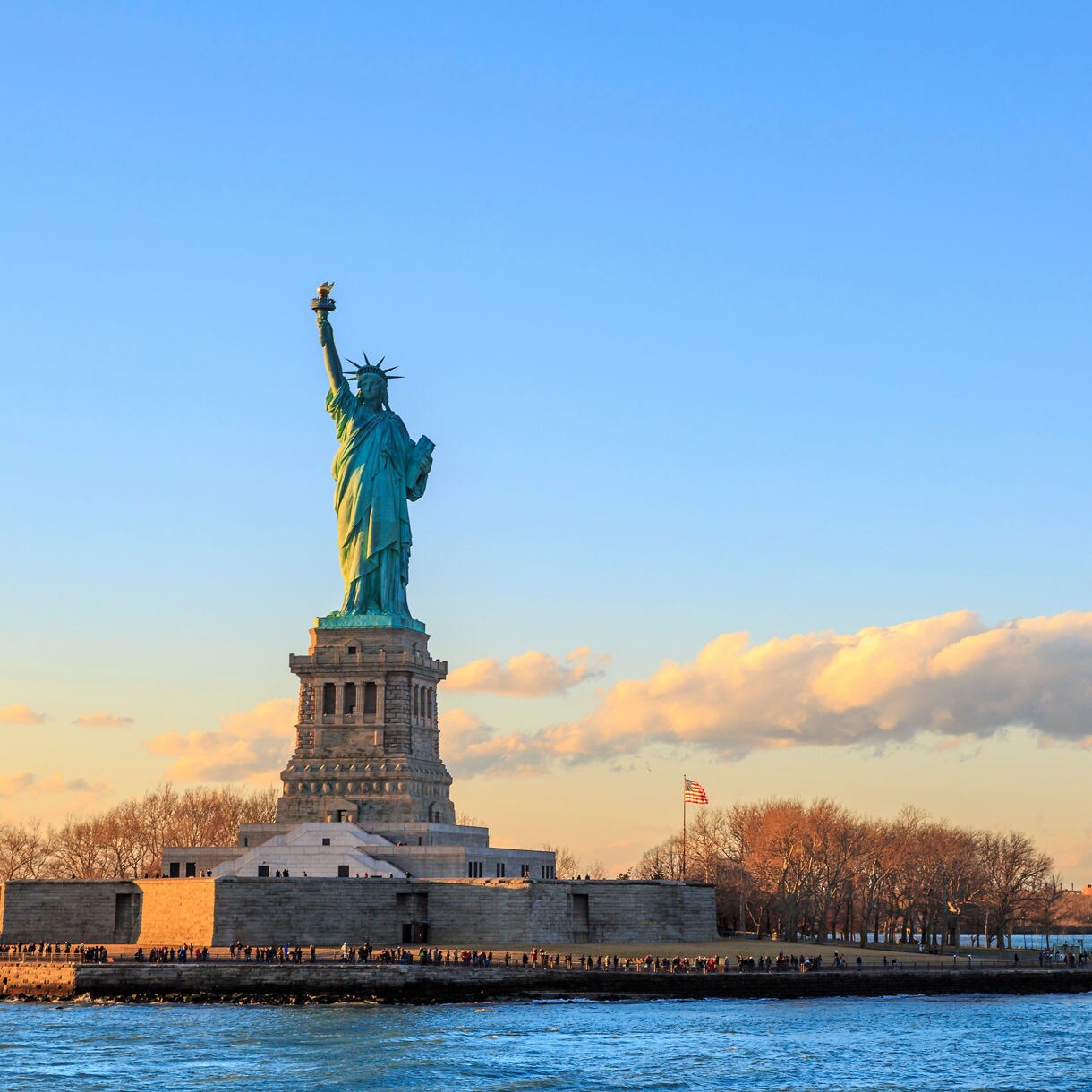 Wide view of the Statue of Liberty rising above its pedestal at sunset, with warm light on the monument, leafless trees along the shoreline and calm harbor water in the foreground.