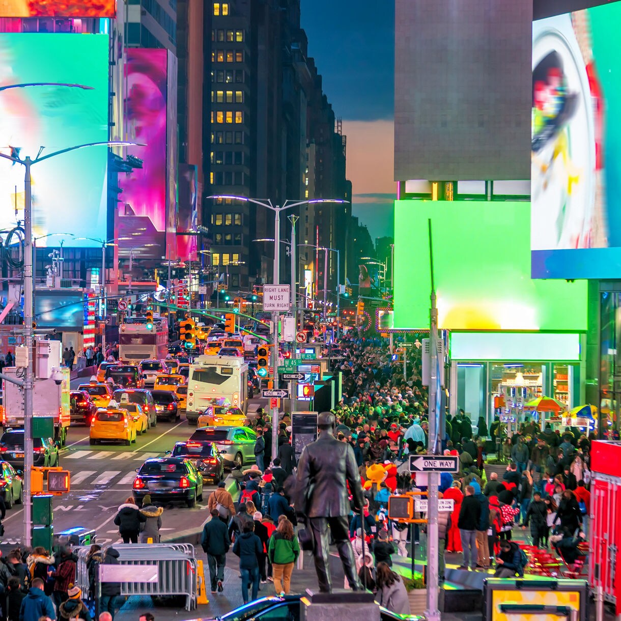 Street-level view of Times Square packed with traffic and pedestrians, surrounded by towering LED billboards glowing in bright colors against the evening sky.