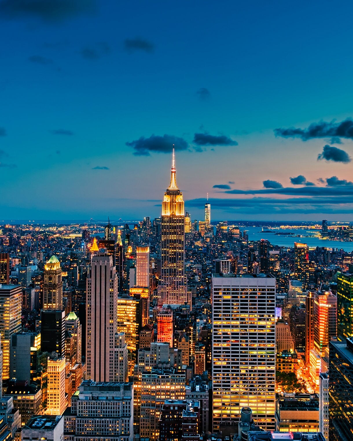 Aerial view of Manhattan at twilight with the Empire State Building glowing brightly among illuminated skyscrapers, set against a deep blue sky fading into sunset colors.