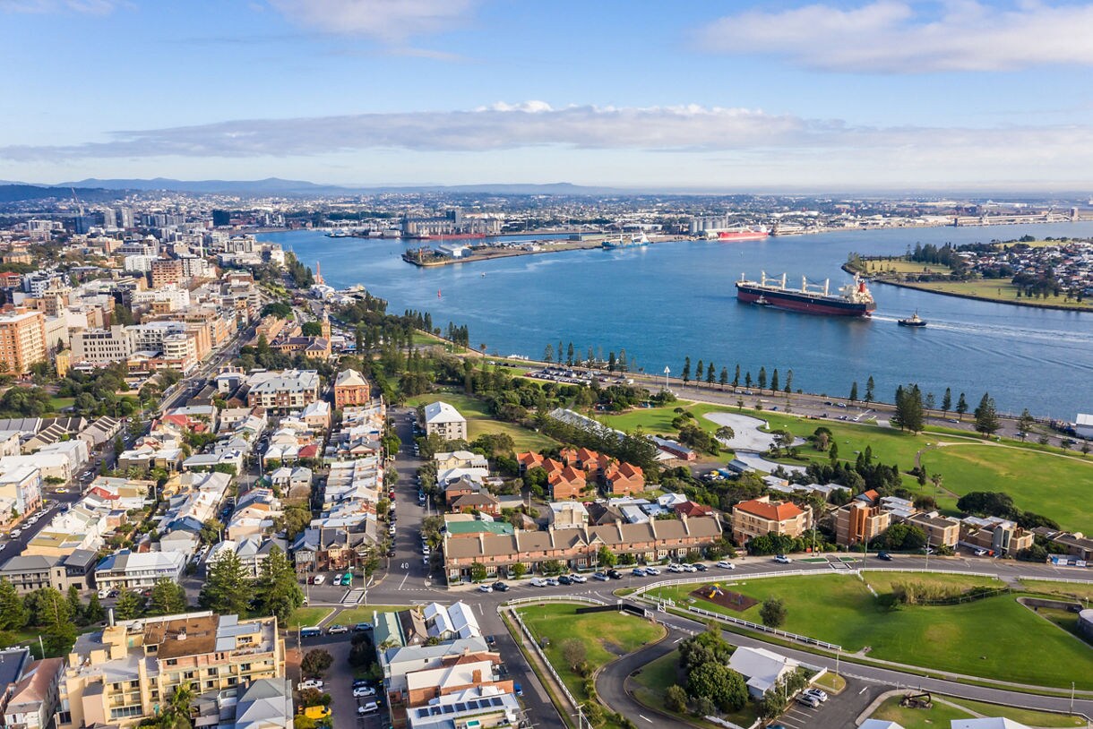 Aerial view of Newcastle, Australia showing residential neighborhoods, green parkland and cargo ships moving through a wide blue harbor.