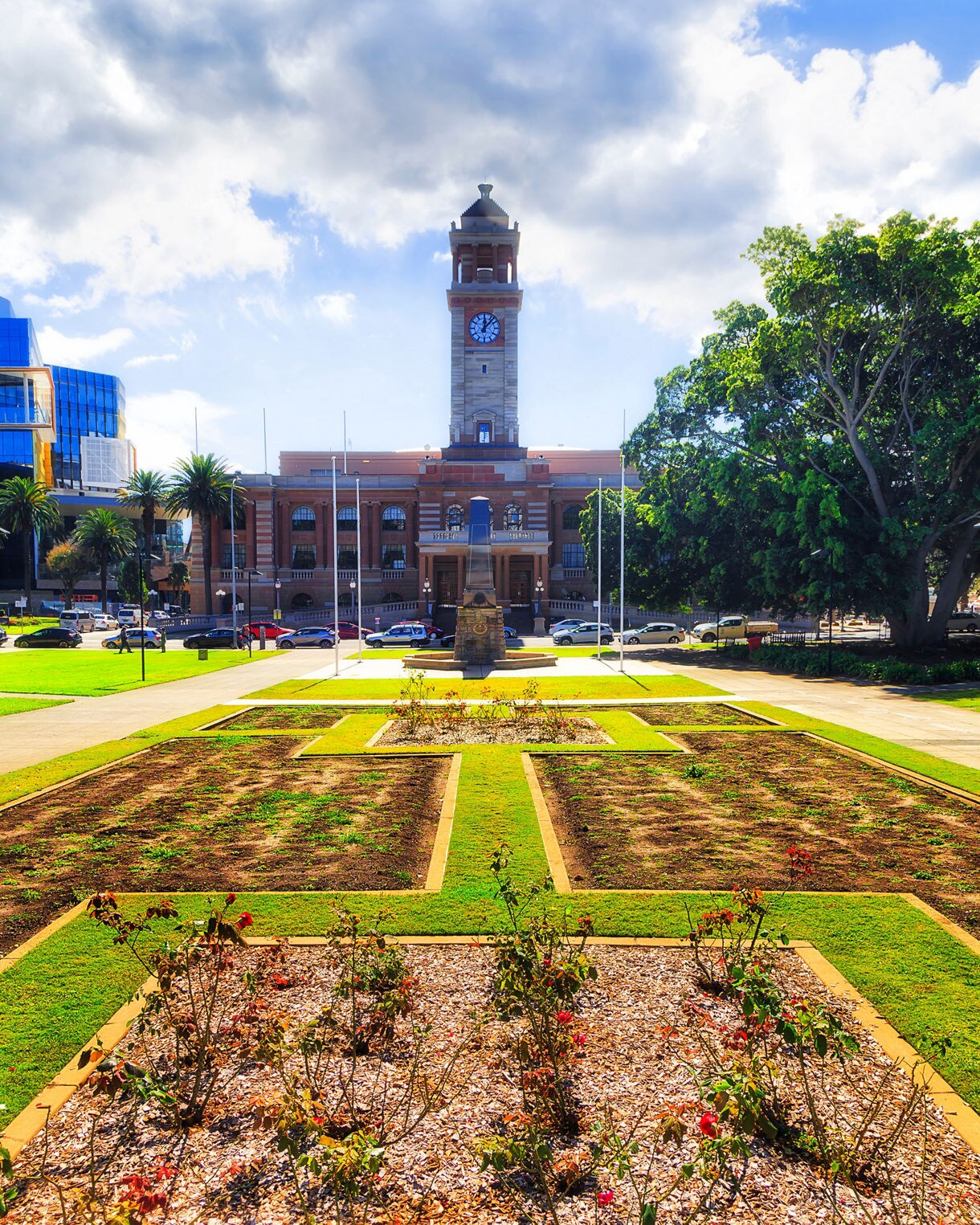 Newcastle Town Hall with its tall clocktower viewed from a garden in Civic Park, surrounded by lawns, trees and city buildings under a partly cloudy sky.