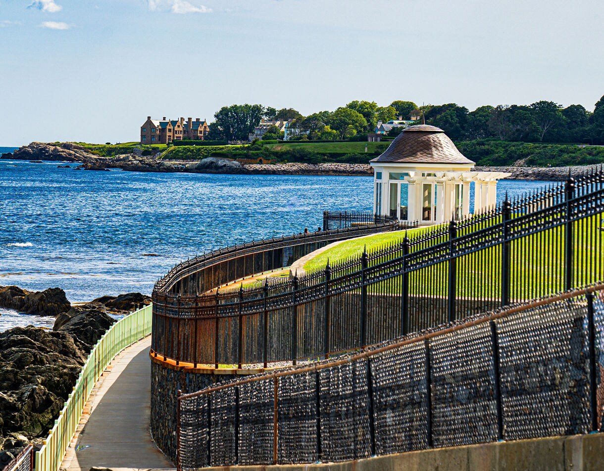 Curving seaside walkway lined with black iron fencing overlooking rocky shore, a small white pavilion and distant mansions under a bright sky.