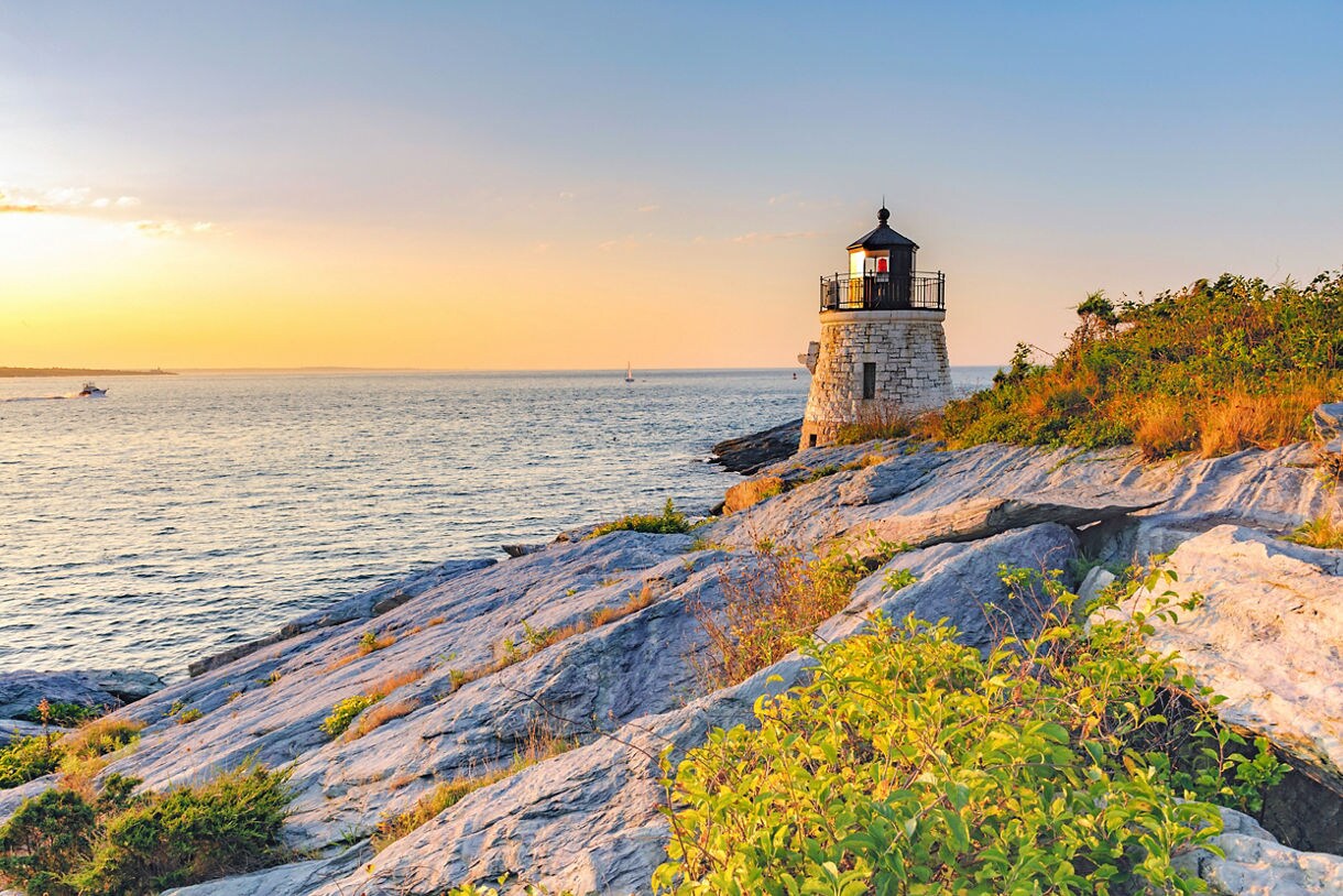 Small round stone lighthouse on rocky shoreline overlooking calm ocean waters at sunset with warm orange light.