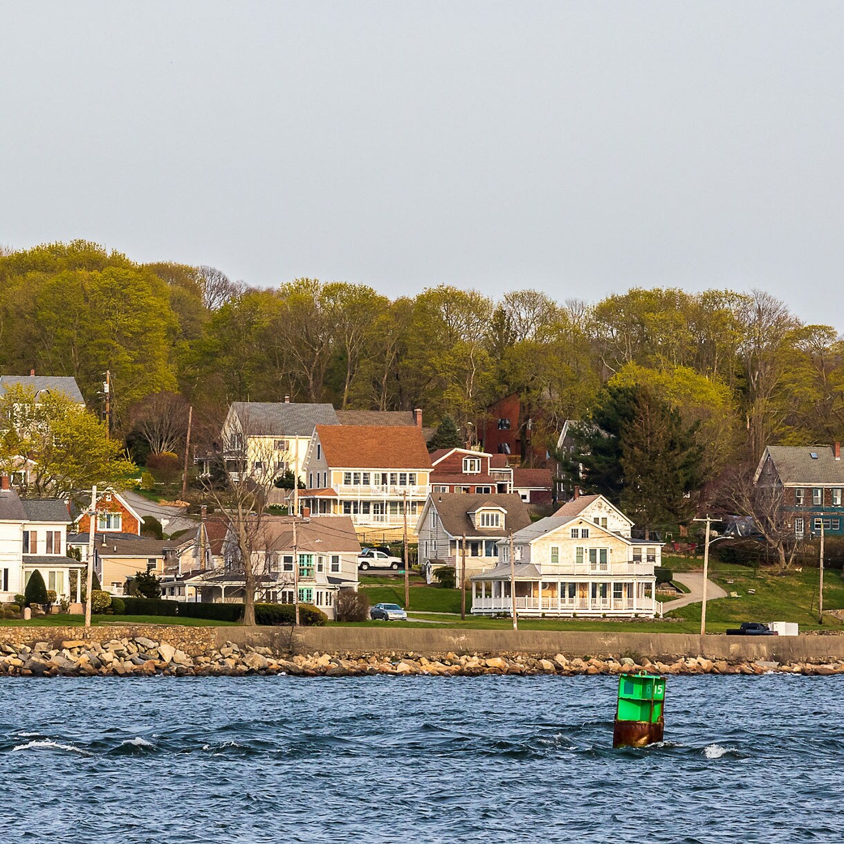 Row of seaside New England houses set above a rocky seawall with spring trees behind and choppy blue water in the foreground.