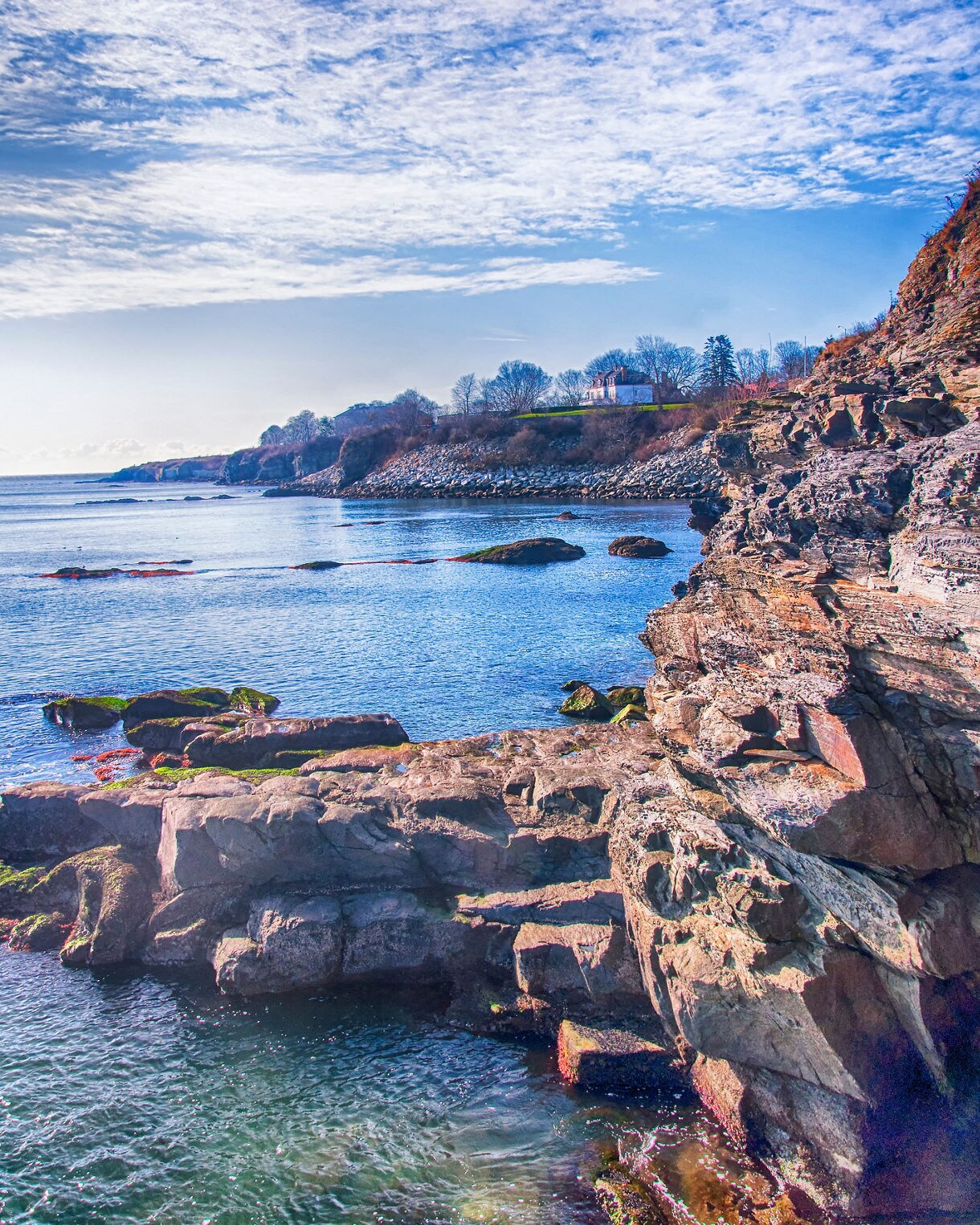 Rocky coastal cliffs overlooking bright blue water with scattered boulders and a distant house set above the shoreline under a wide, cloud-streaked sky.
