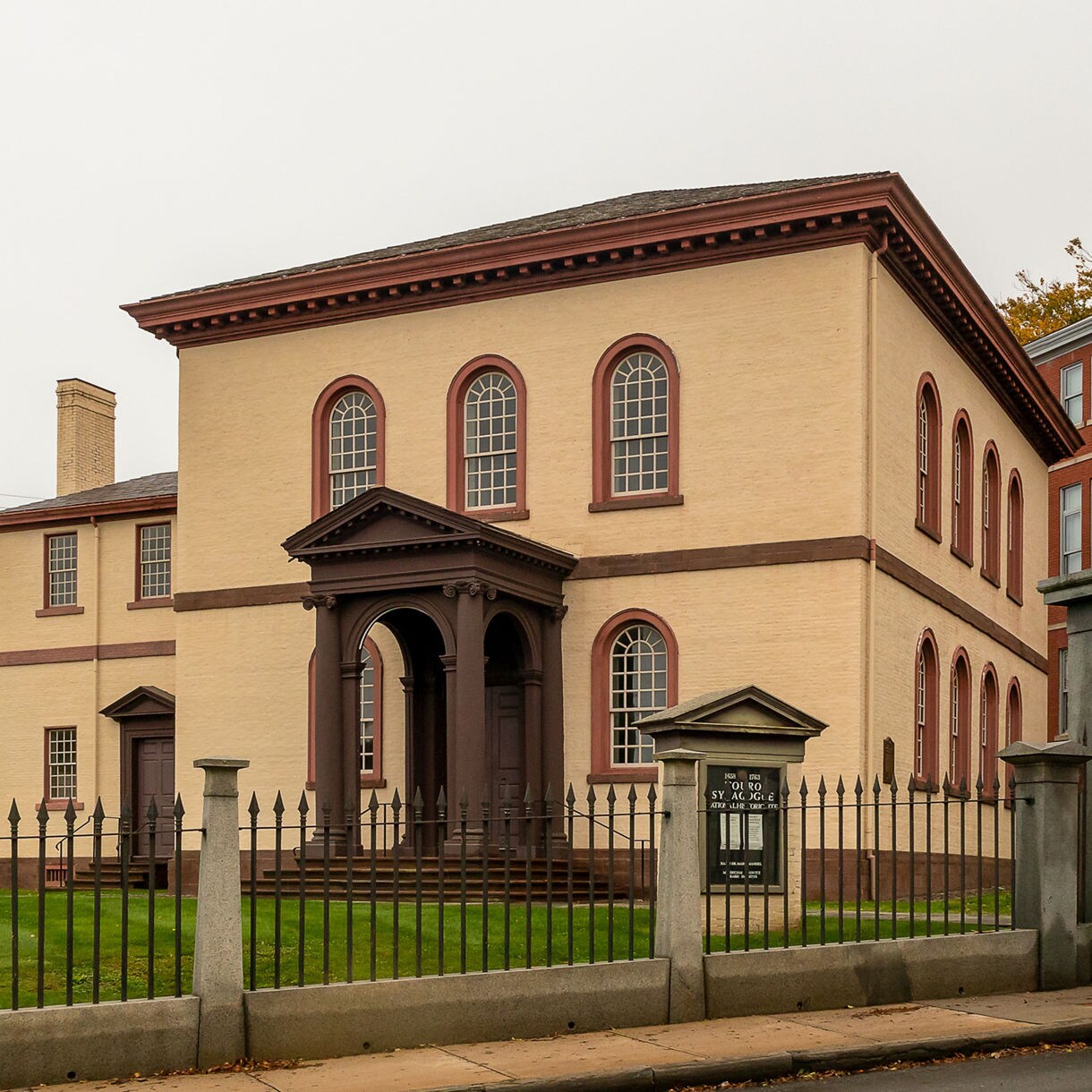 Exterior view of the Touro Synagogue in Newport, showing its cream brick facade, arched windows and gated entrance surrounded by historic buildings.