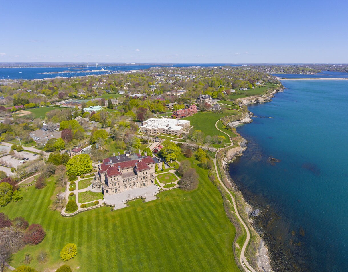 Aerial view of Newport’s cliffside with a large historic mansion, sweeping green lawns and a curving coastal path beside calm blue ocean.