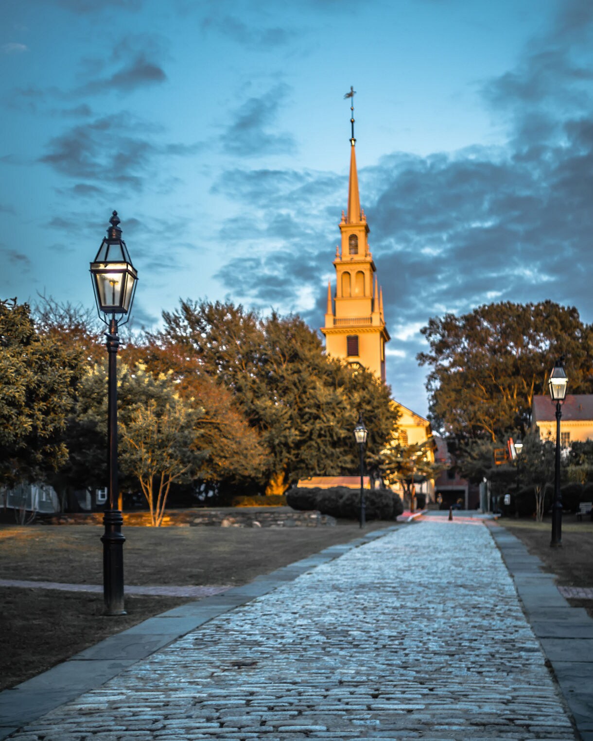 Cobblestone path lined with vintage street lamps leading to a tall church steeple illuminated at sunset with trees on both sides.