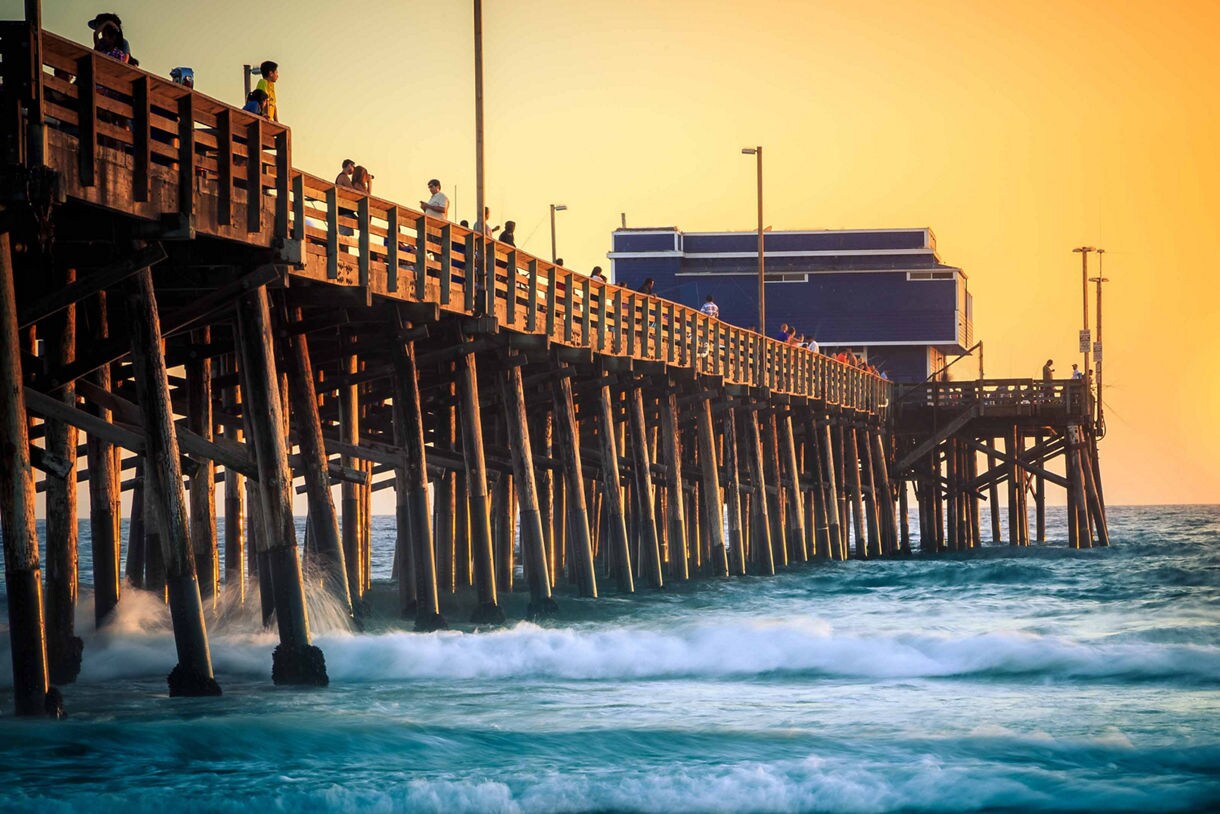 Newport Beach Pier at sunset, with golden light reflecting off the wooden structure as waves crash against its pillars.