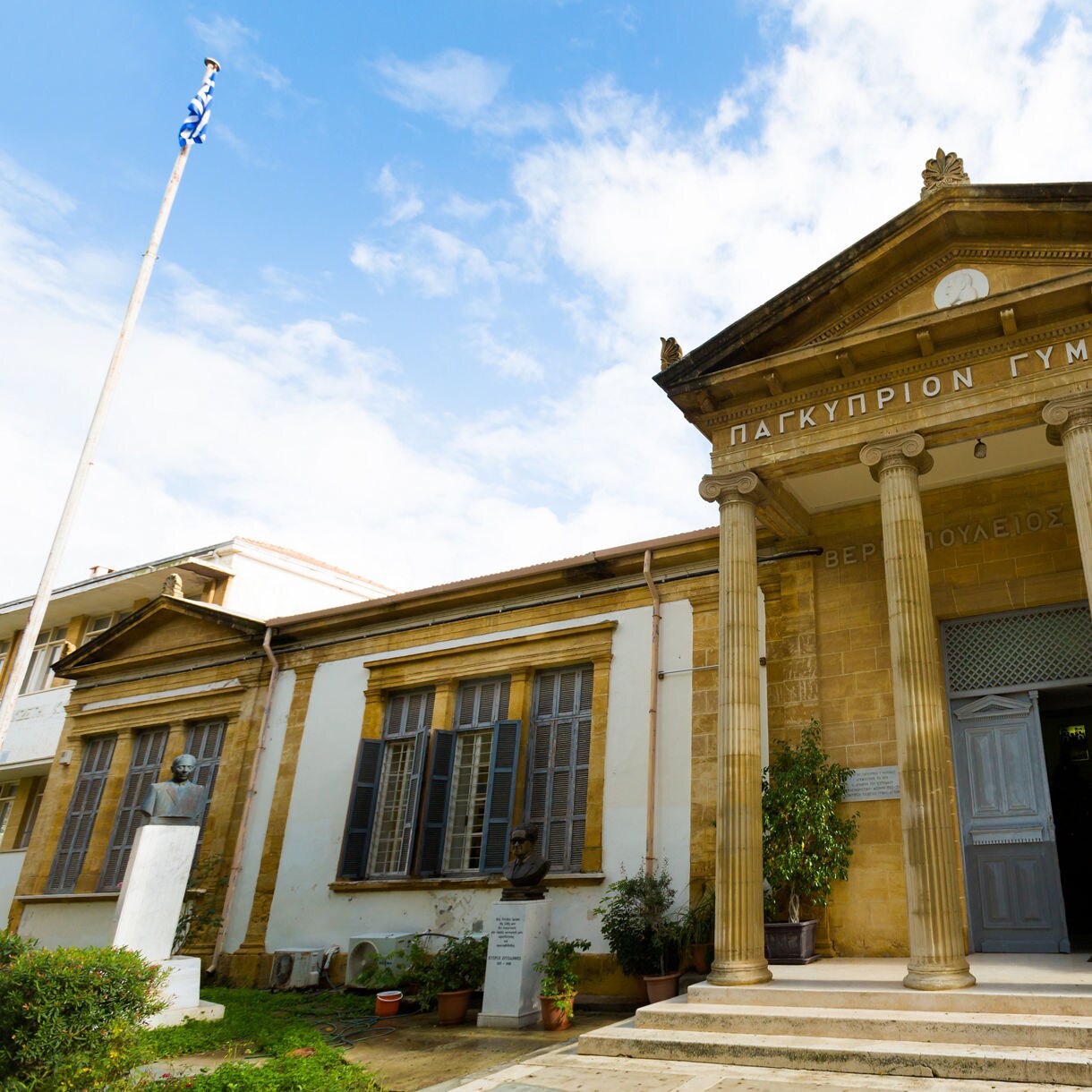 Historic neoclassical school building in Nicosia with tall columns, stone walls and a triangular pediment, featuring a Greek flag and statues near the entrance.