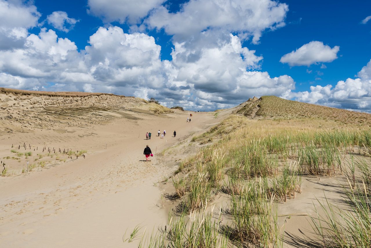 Tourists walking along the vast sandy dunes of Nida on the Curonian Spit in Lithuania, under a bright blue sky filled with white clouds.