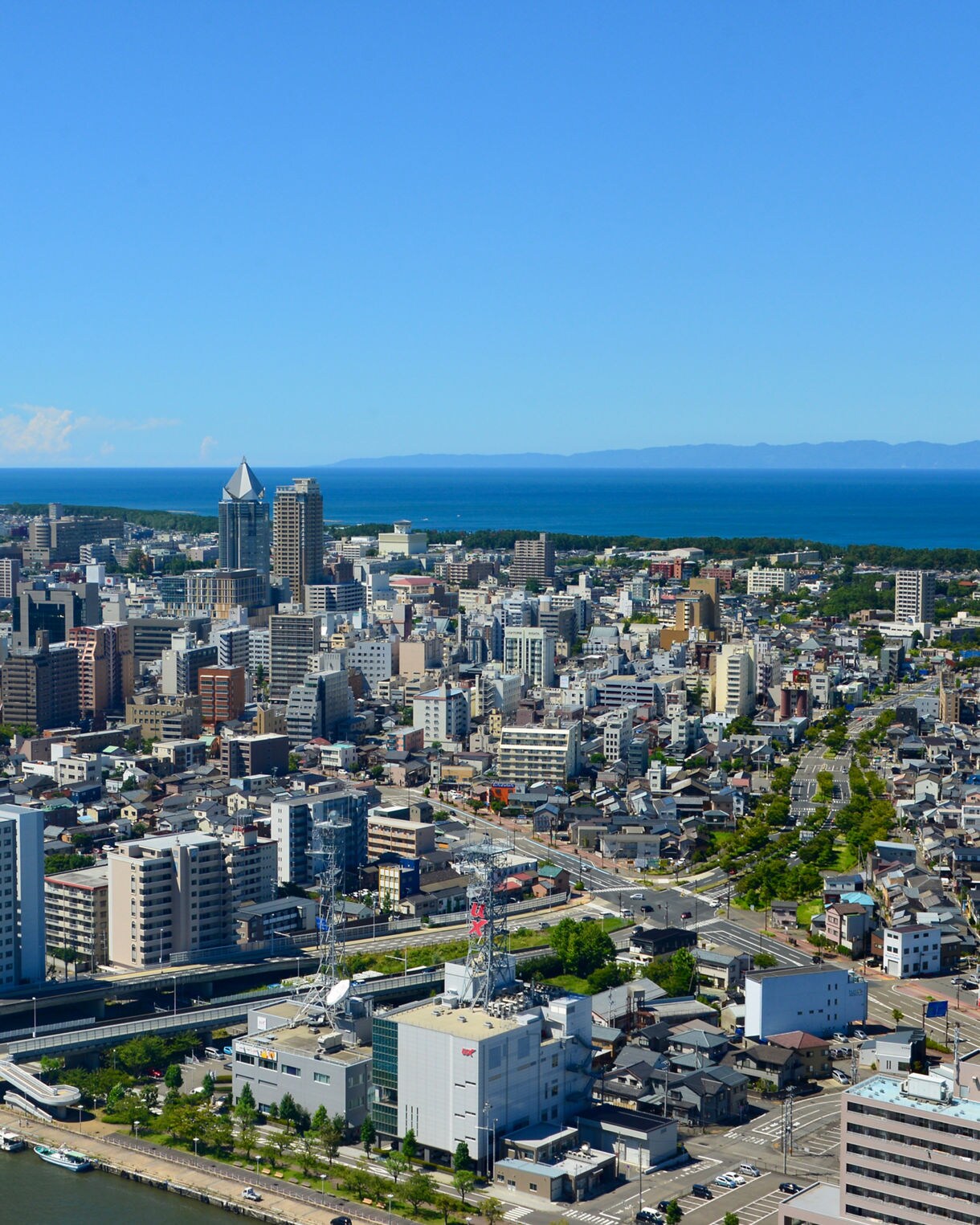 Aerial view of Niigata, Japan, showing dense clusters of high-rise buildings, residential streets and riverside bridges with the Sea of Japan and distant mountains on the horizon.