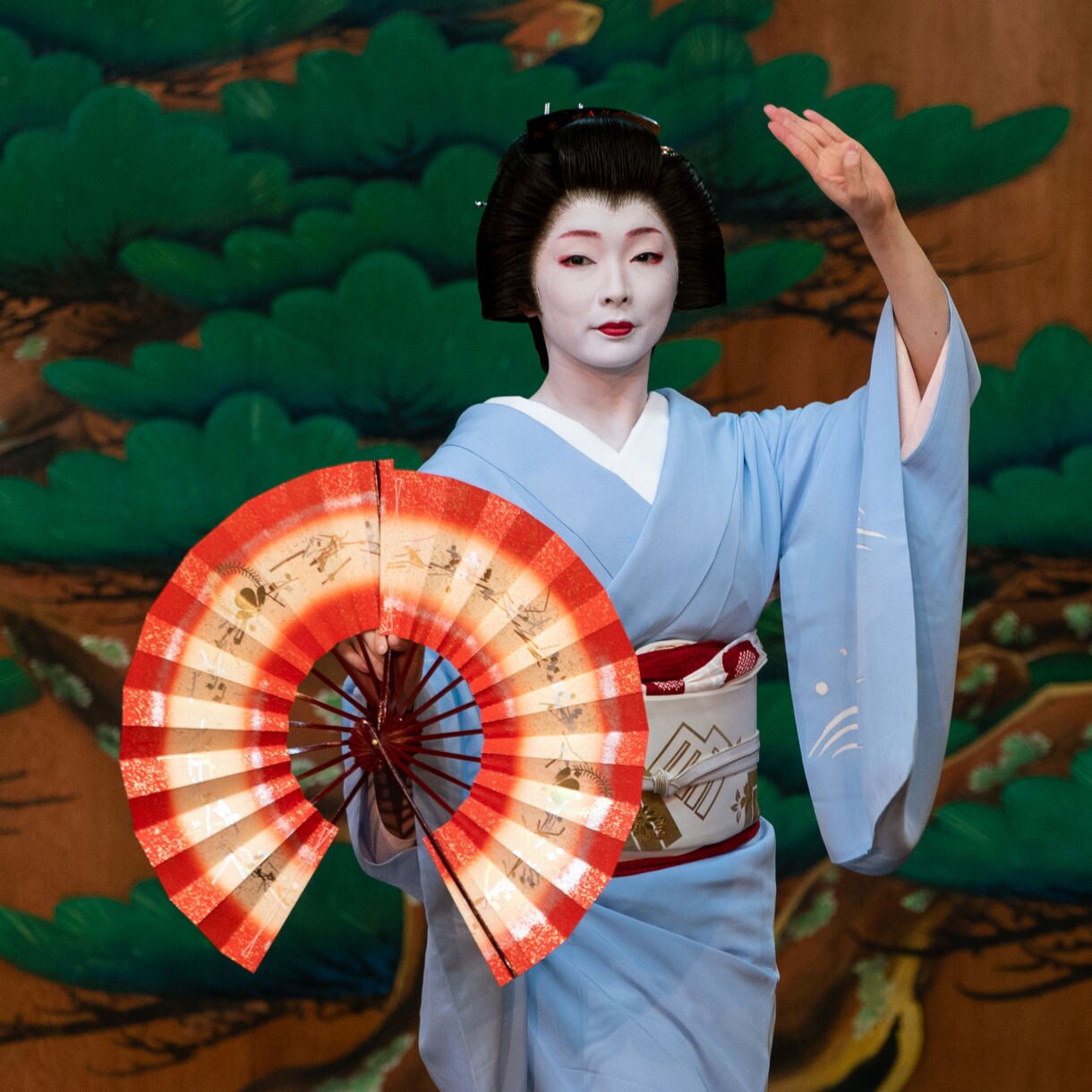 A geigi performer in a pale blue kimono dances on stage while holding a red and white fan, set against a backdrop painted with green pine branches.