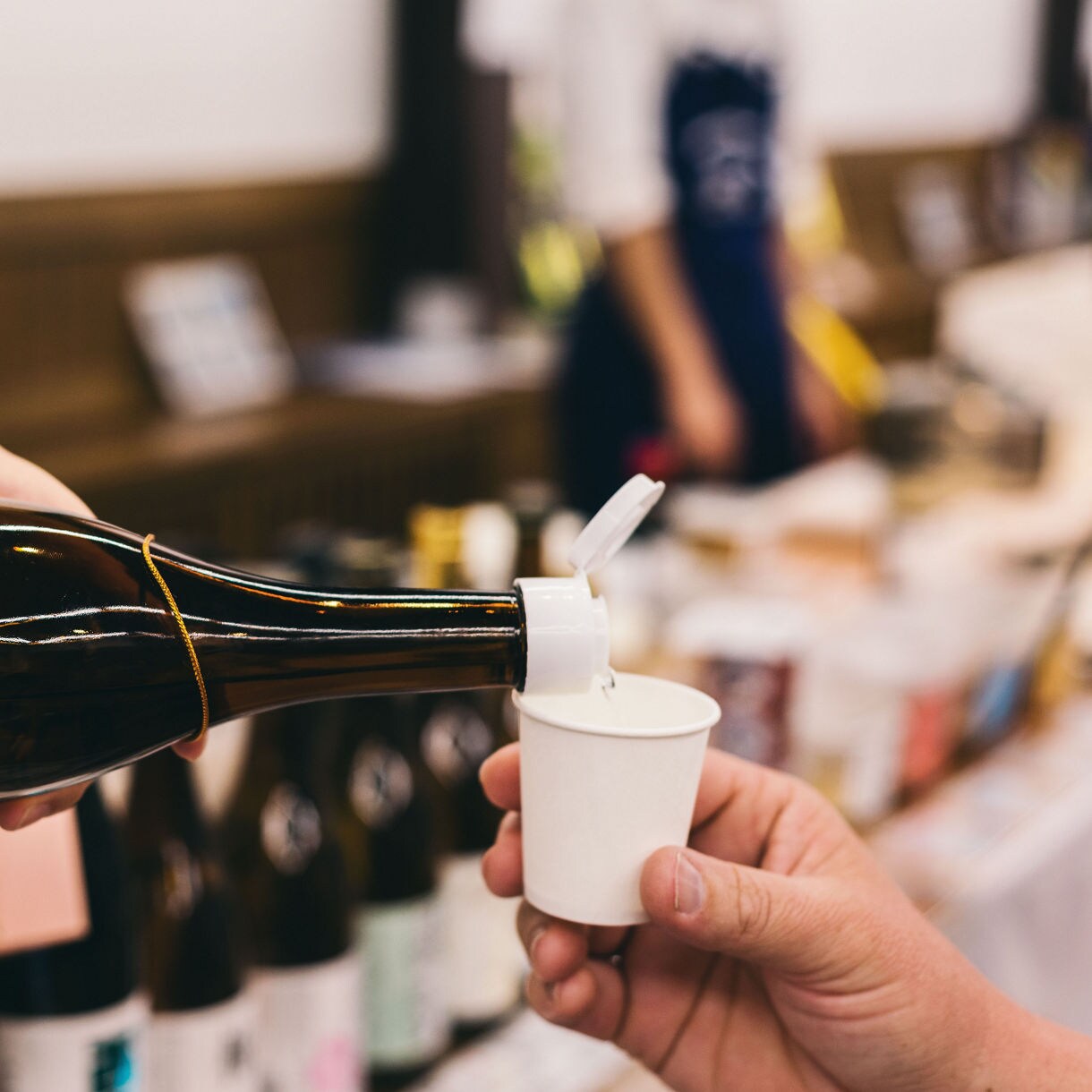Close-up of a person pouring sake from a brown bottle into a small white paper cup at a tasting counter, with blurred bottles and people in the background.