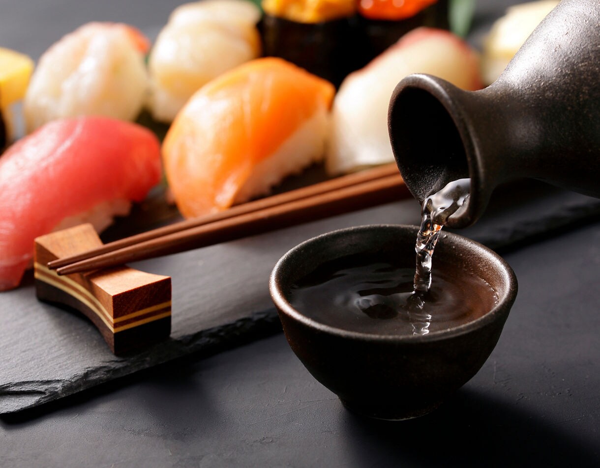 Close-up of assorted sushi pieces on a slate board with chopsticks resting beside them, while clear sake is being poured from a ceramic flask into a small matching cup in the foreground.