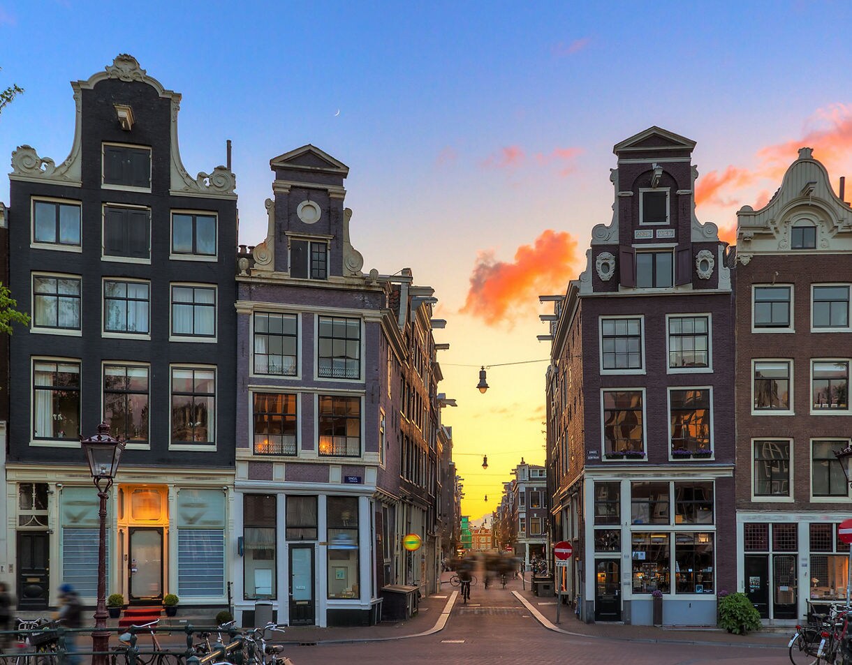 Sunset view of a narrow street in Amsterdam’s Nine Streets district, lined with historic Dutch canal houses featuring ornate gables and large windows, with soft evening light illuminating the buildings.