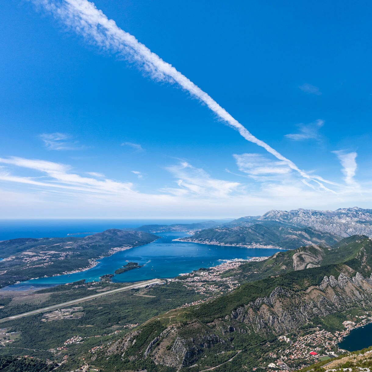 Aerial view from Njeguši overlooking the Bay of Kotor in Montenegro, with deep blue waters, cruise ships, coastal towns and dramatic mountain ridges under a bright sky.