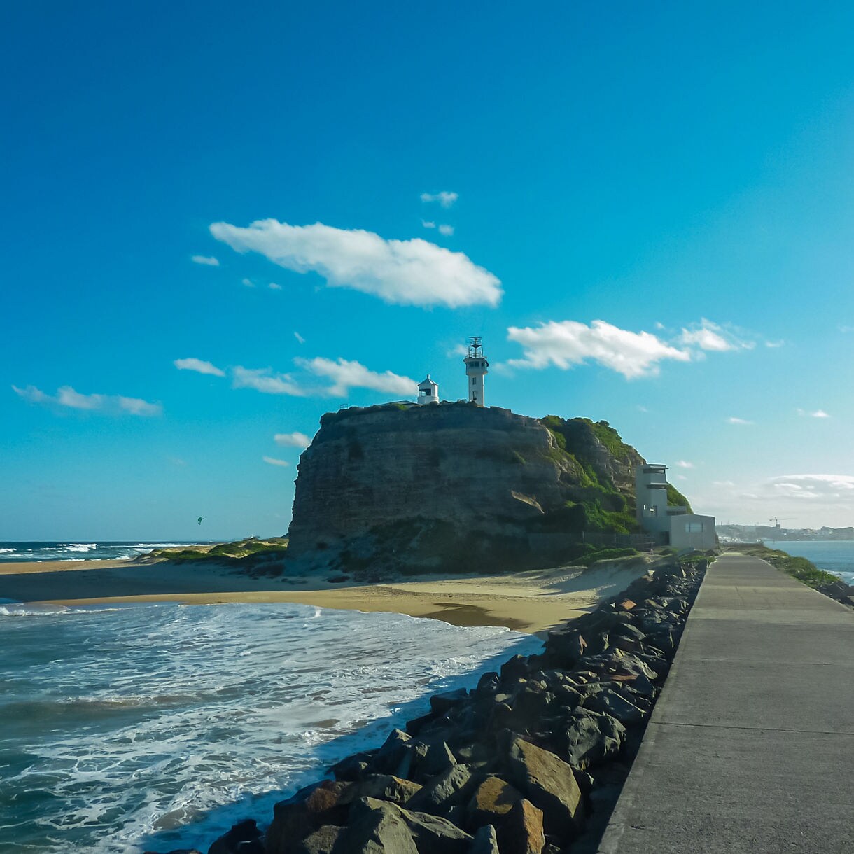 Nobbys Lighthouse perched on a rocky headland, with waves rolling onto a sandy beach and a long walkway leading toward the lighthouse.