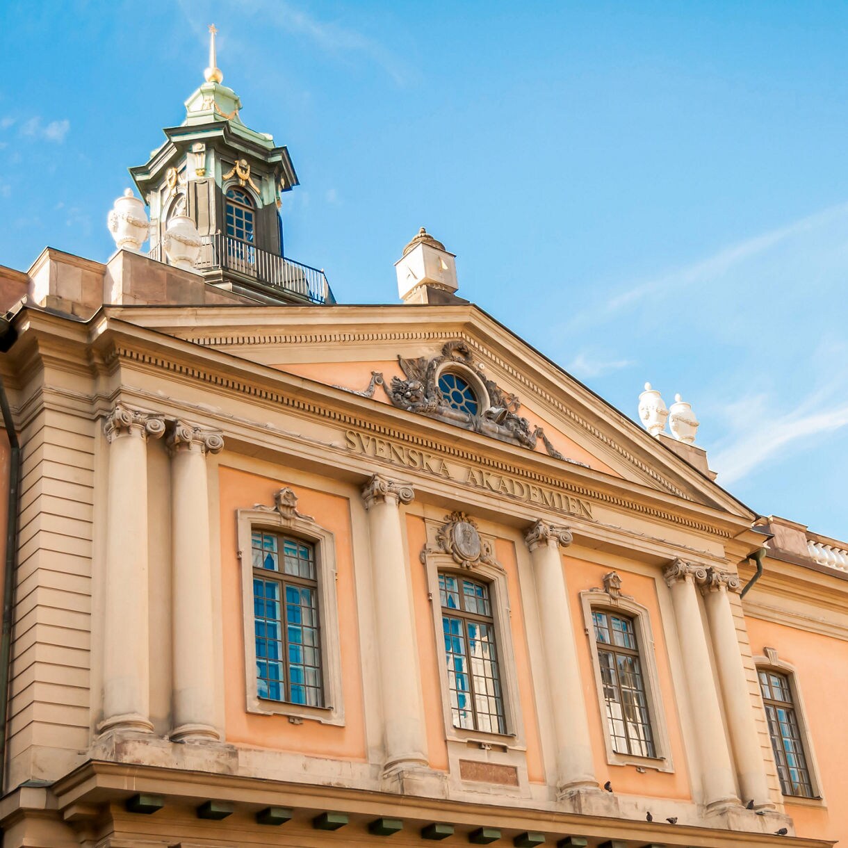 The Nobel Prize Museum building in Stockholm, featuring neoclassical architecture with tall columns, arched windows and a central clock under a blue sky.