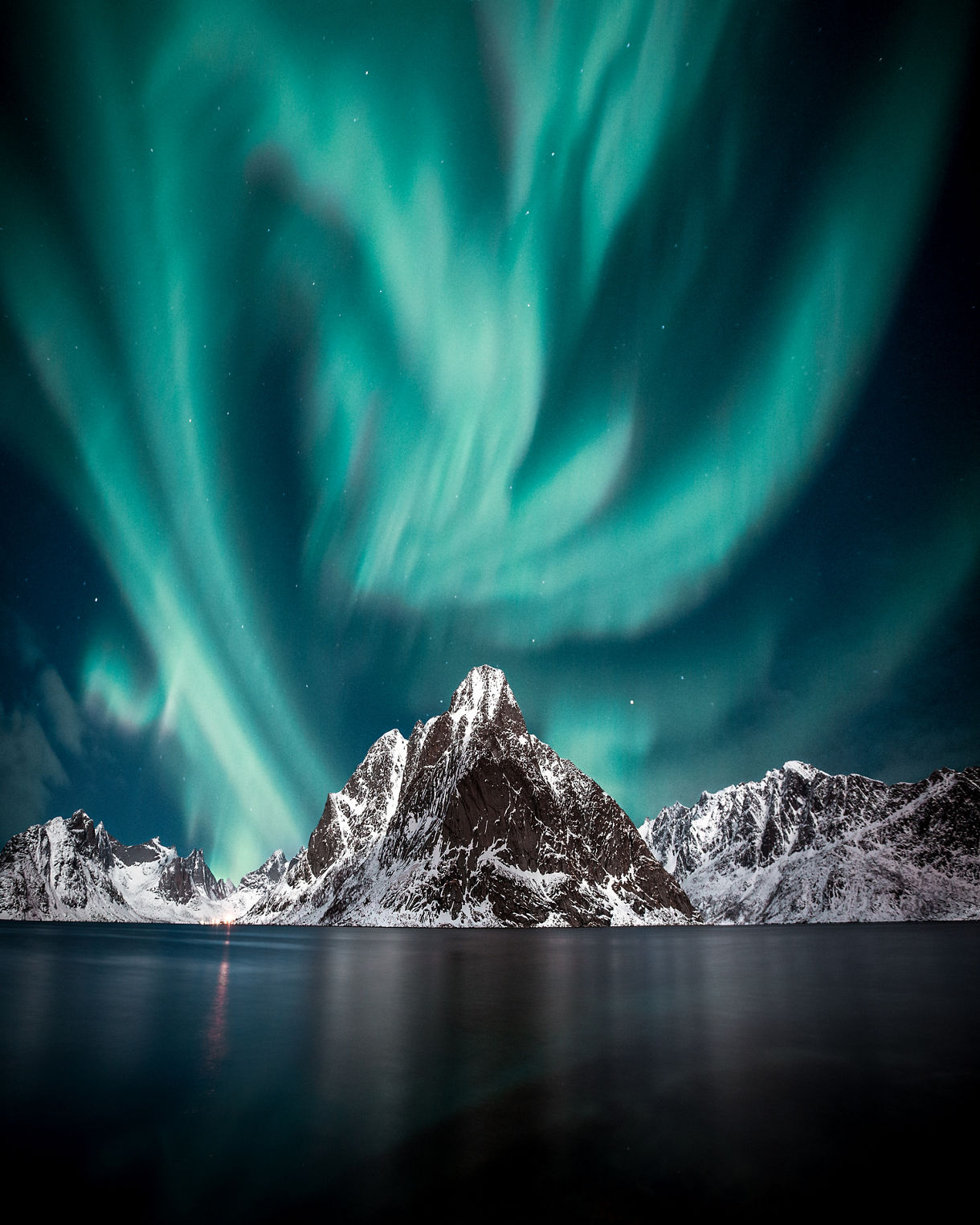 Green aurora borealis swirls above jagged snow-covered mountains reflected in calm Arctic waters in Lofoten, Norway.