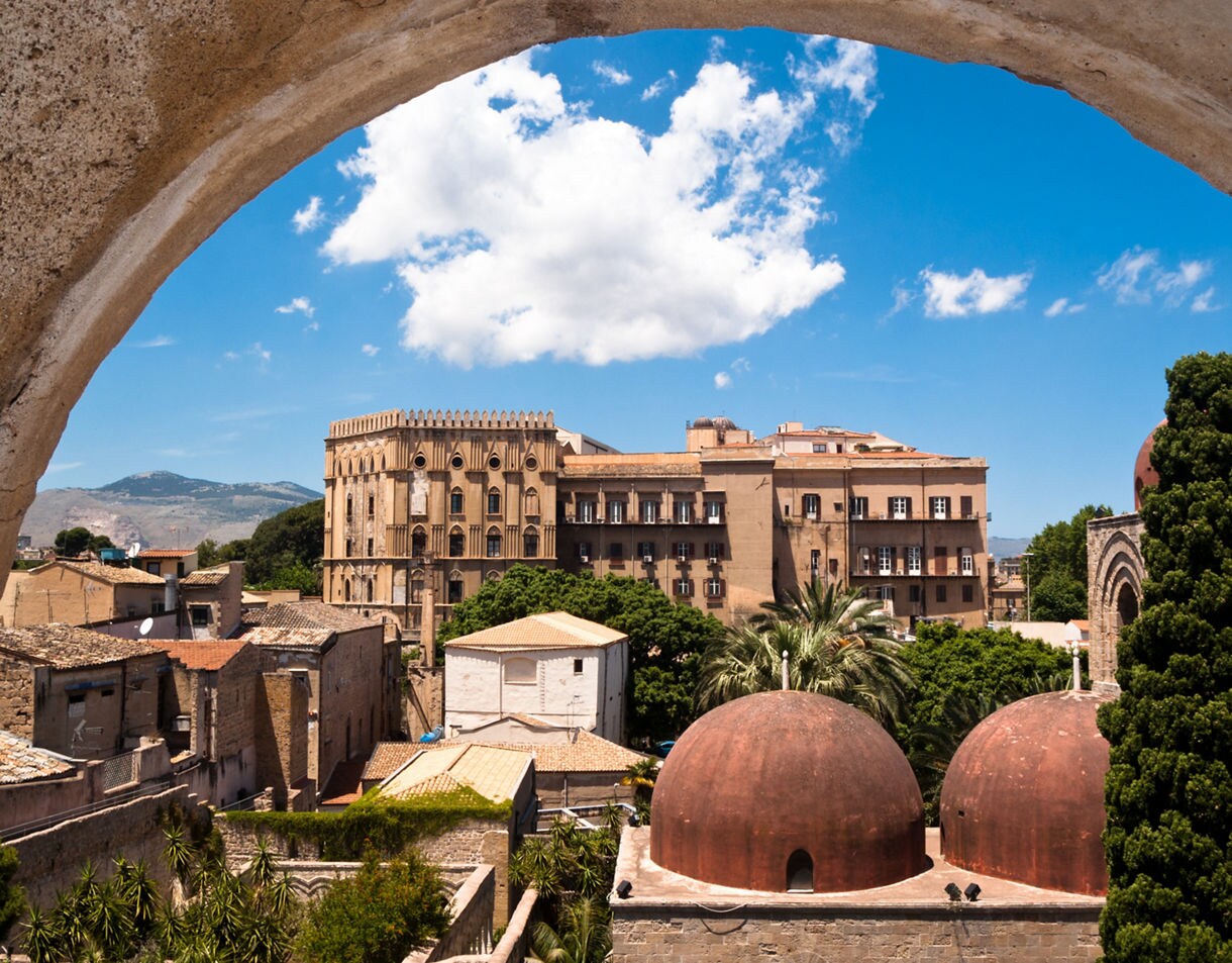 View of Palermo’s historic center featuring terracotta-domed rooftops, palm trees and the Norman Palace in the background framed by a stone arch.