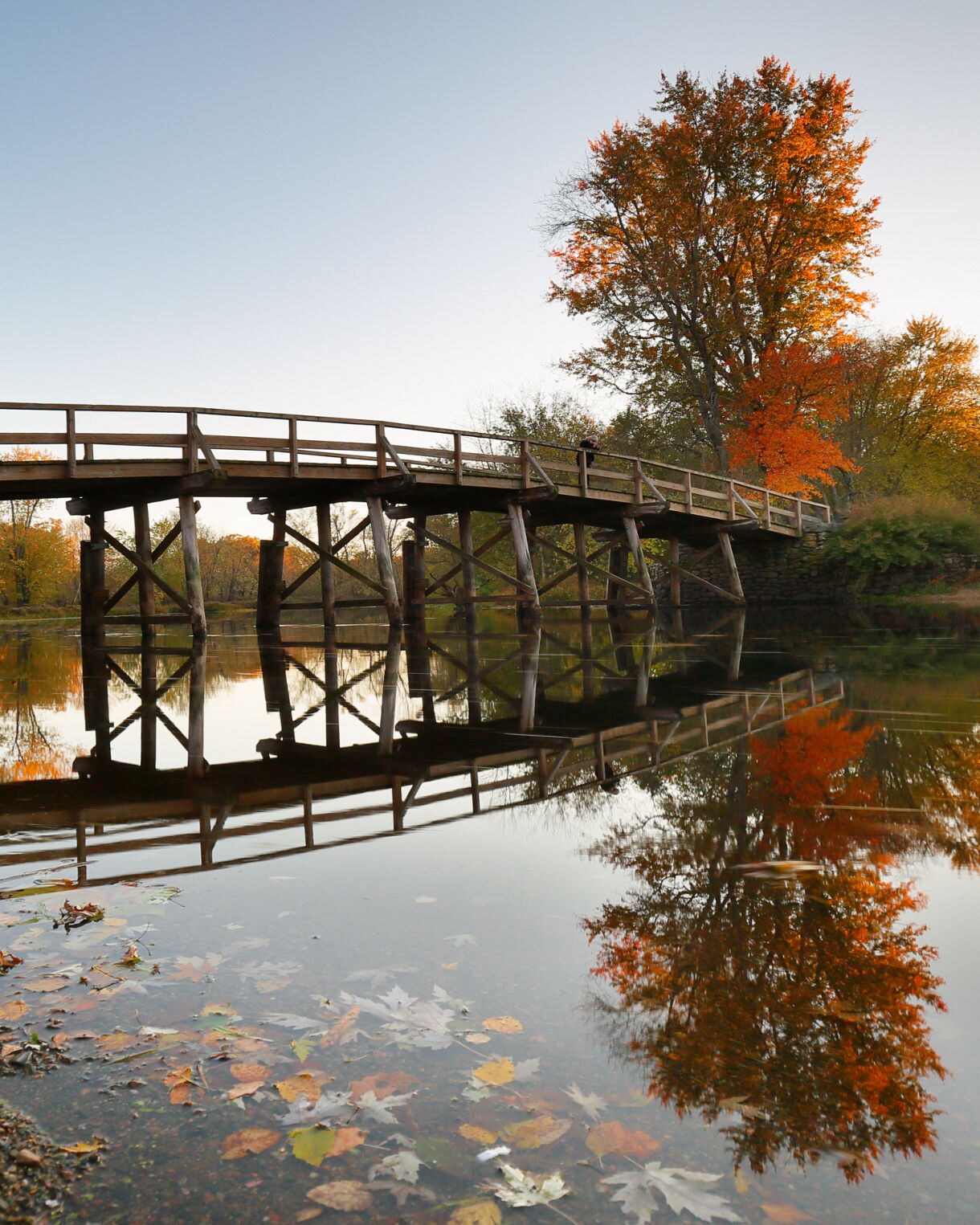 Wooden bridge spanning a calm river in Concord, Massachusetts, with fall trees and their reflections mirrored in the water.