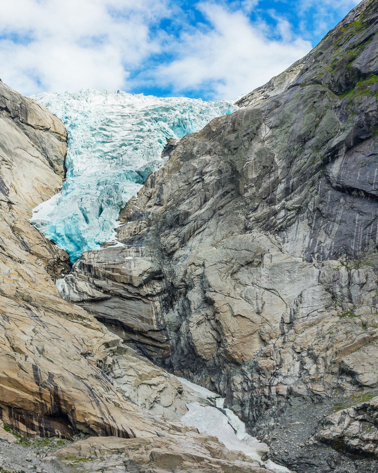 Briksdal Glacier in Norway with bright blue ice descending through a steep rocky valley under a partly cloudy sky.