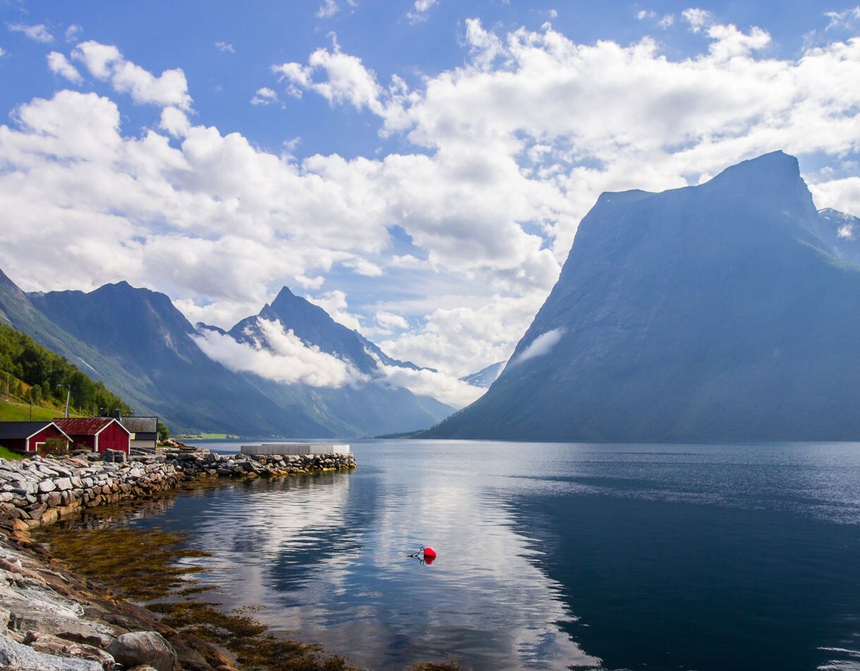 Red wooden cabins along a rocky shoreline on Norway’s Hjørundfjord with towering mountains, drifting clouds and calm reflective water.