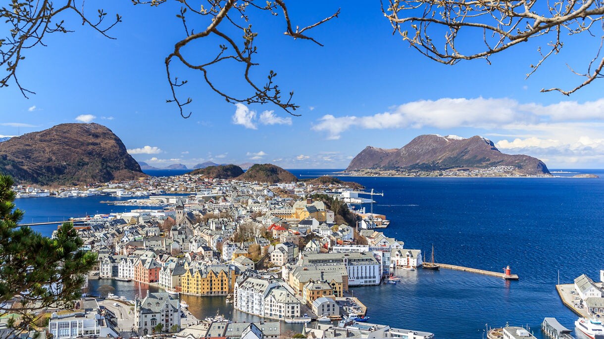 View of Ålesund Norway featuring pastel waterfront buildings, deep blue harbor waters and surrounding mountains under a clear sky framed by tree branches.