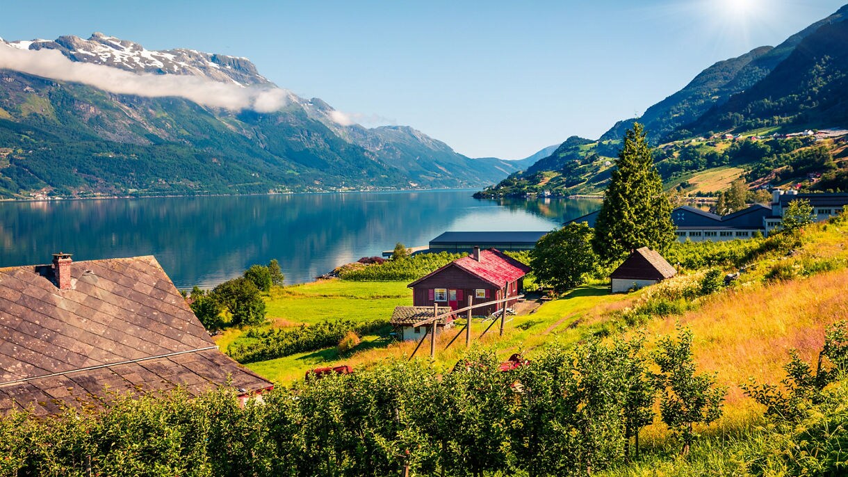 Sunny view of Hardangerfjord Norway featuring red wooden houses, green orchards, calm reflective fjord waters and snow-dusted mountains in the distance.