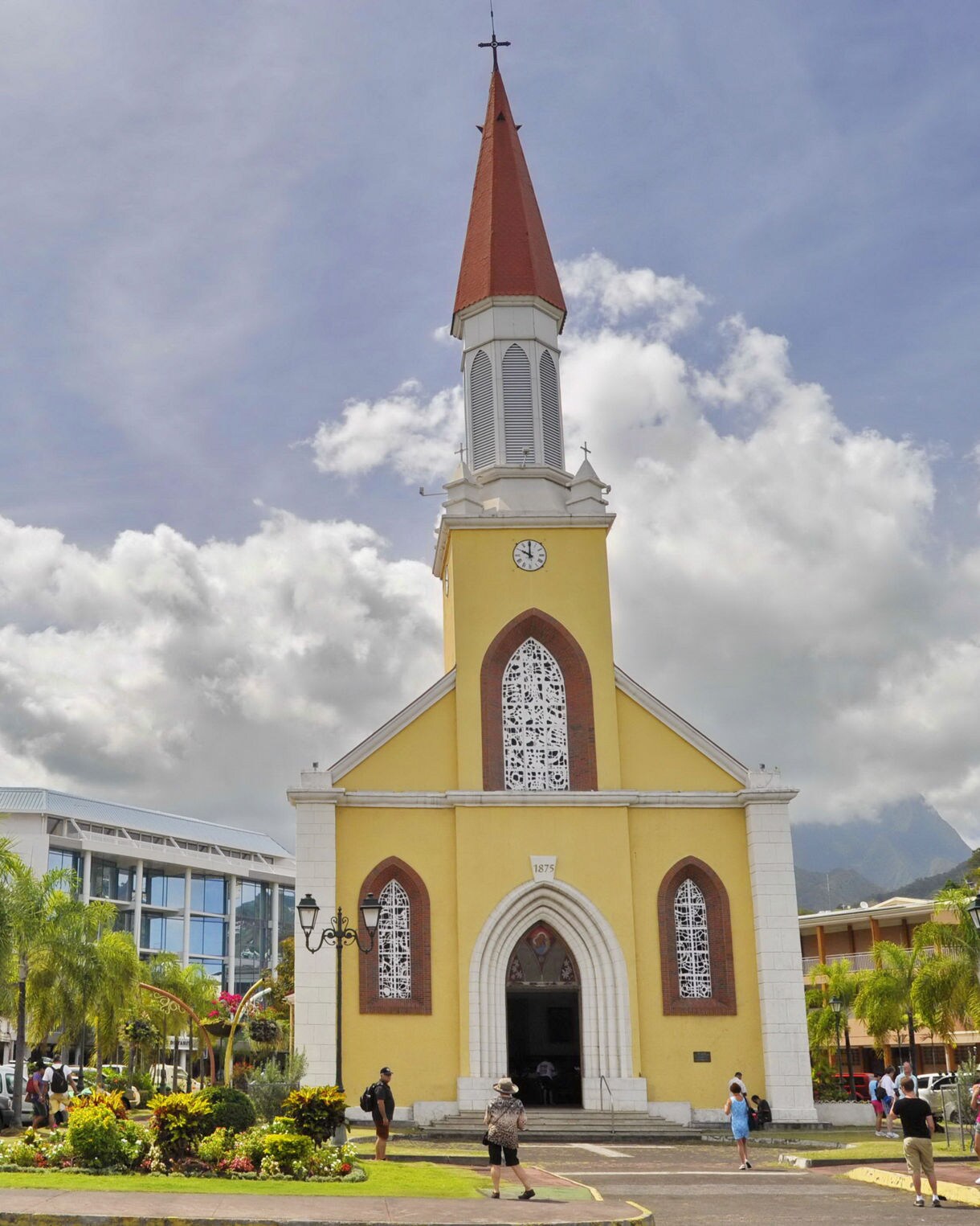Yellow Notre Dame Cathedral in Papeete with a tall red steeple, arched white-trimmed windows and people walking through a landscaped plaza surrounded by modern buildings.